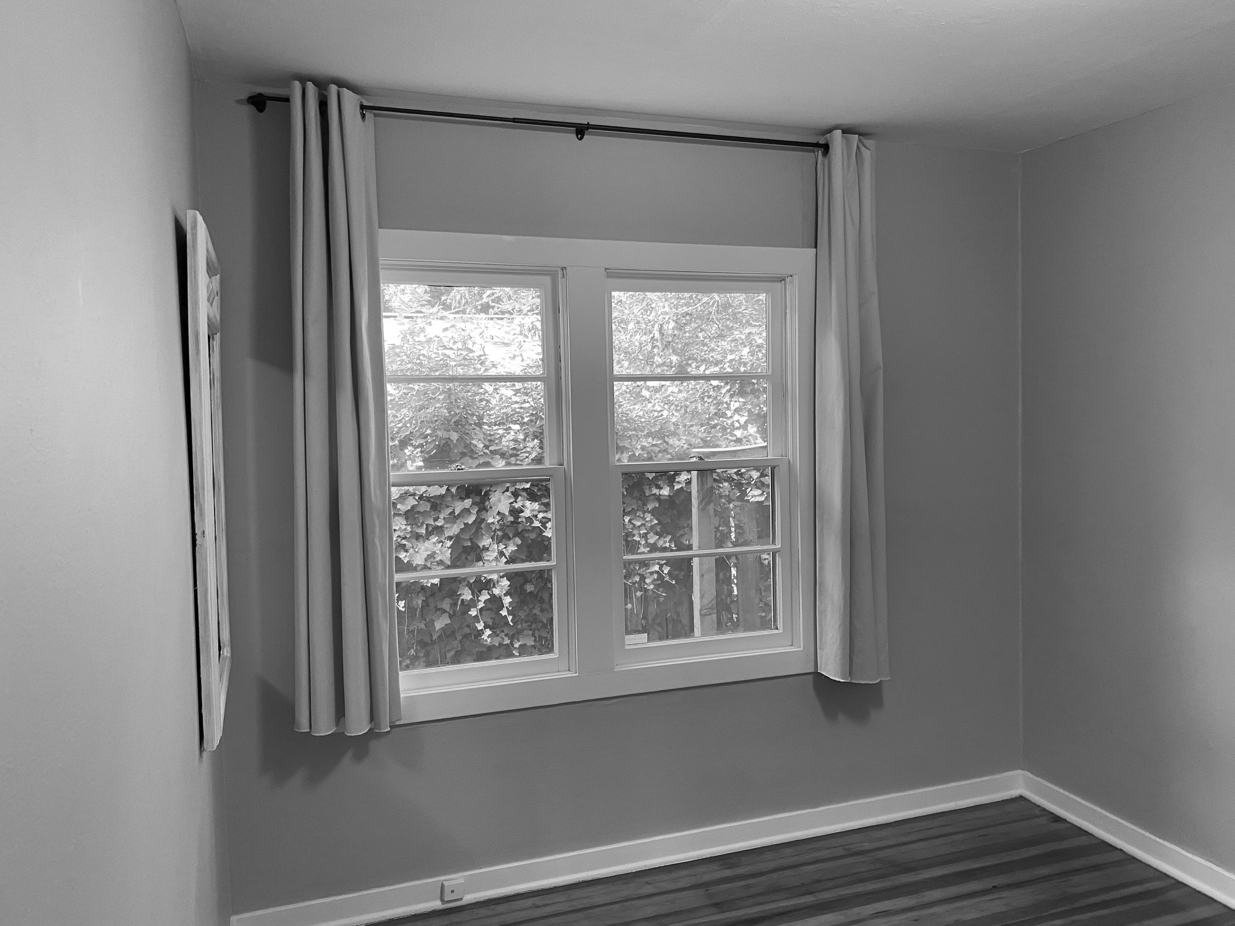 Black and white photo of a room with a large window and curtains. Outside view shows foliage. Minimalist decor.