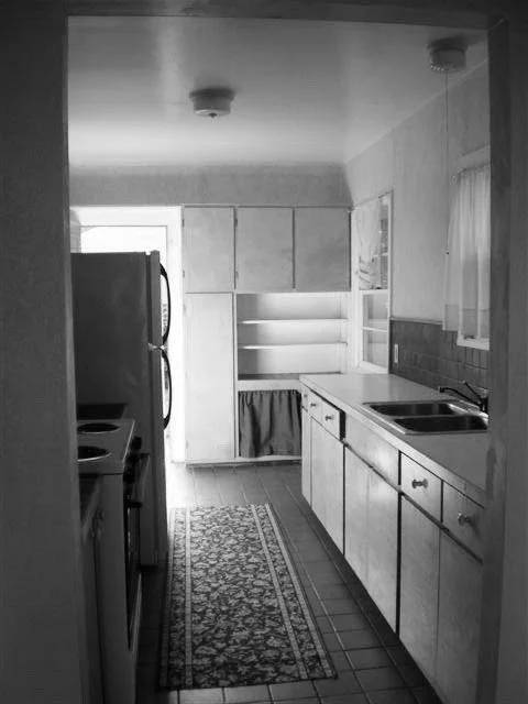 Black and white photo of a narrow kitchen with cabinets, a stove, a refrigerator, and a sink, featuring a tiled floor and a patterned runner rug.