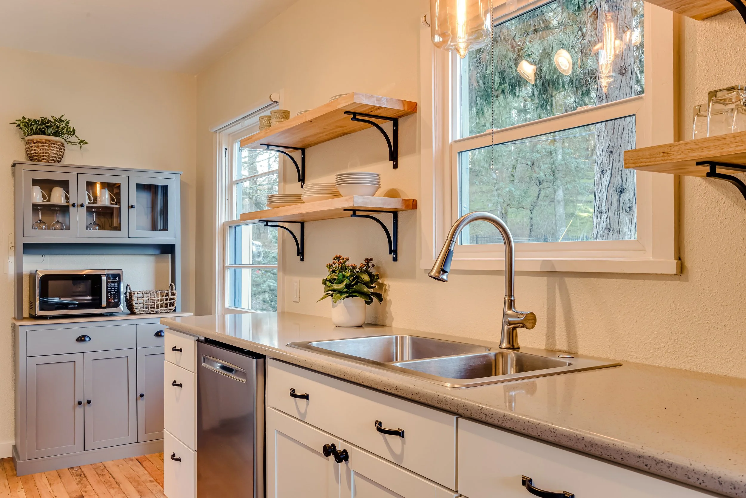 Modern kitchen interior with white cabinets, stainless steel sink, window, shelves with dishes, and a microwave on a gray cabinet.