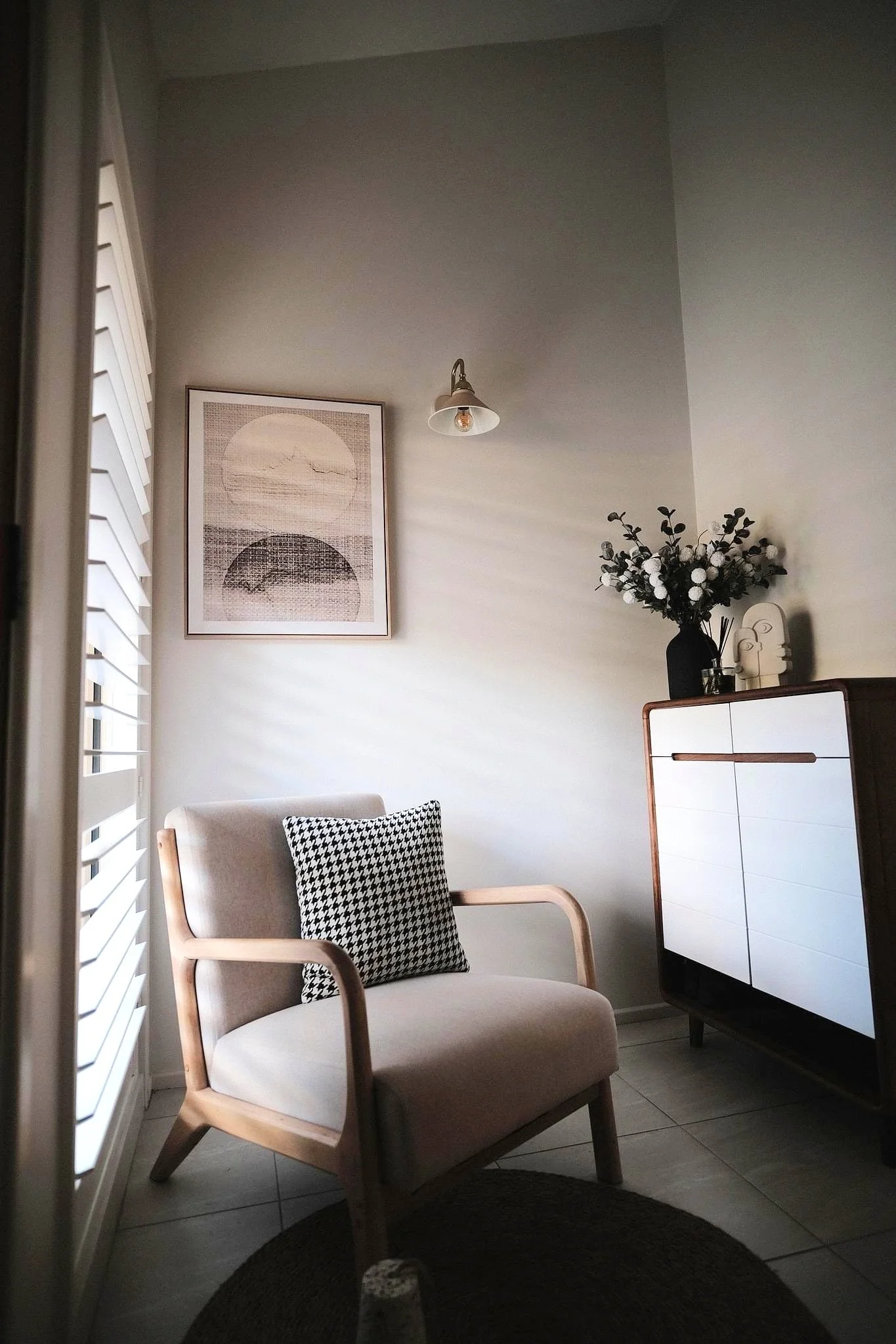 A cozy corner of a modern living room with a beige armchair, black and white patterned pillow, a white and wood cabinet with decorative items, a framed abstract art piece, a black vase with white flowers, and a wall-mounted light, illuminated by natu