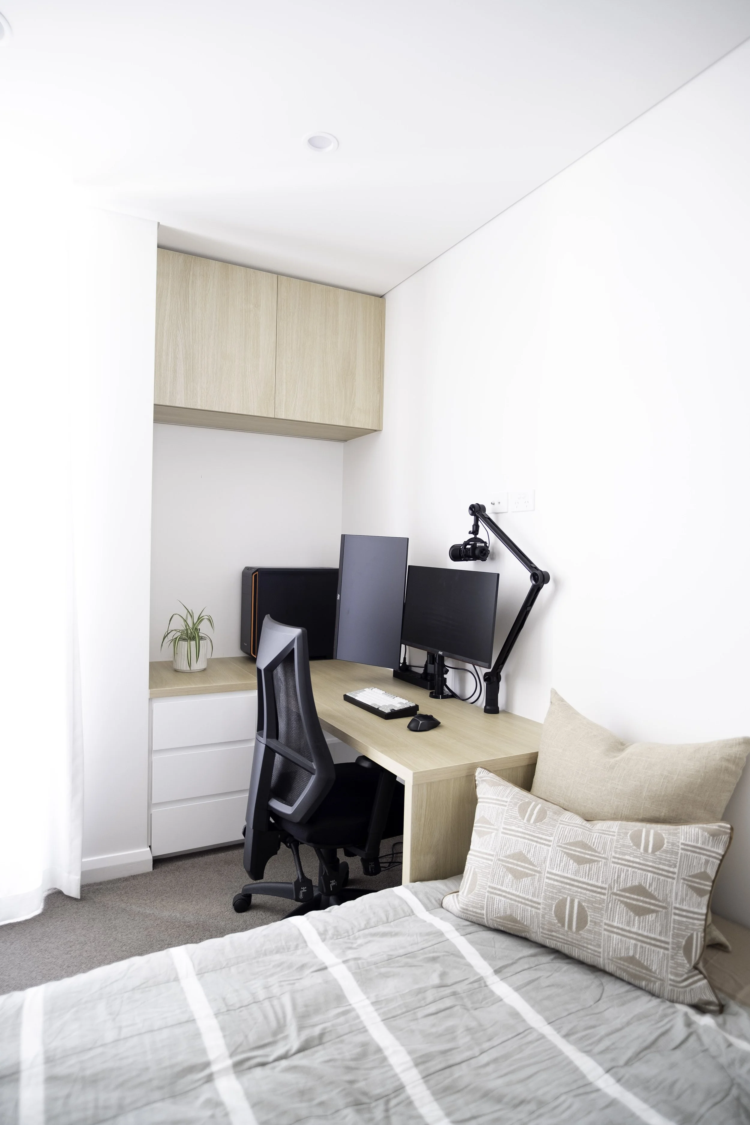 A bedroom with a white bed, beige pillows, a wooden closet, and a white desk with two computer monitors, a keyboard, a mouse, a camera on an adjustable arm, a computer tower, and a small potted plant.