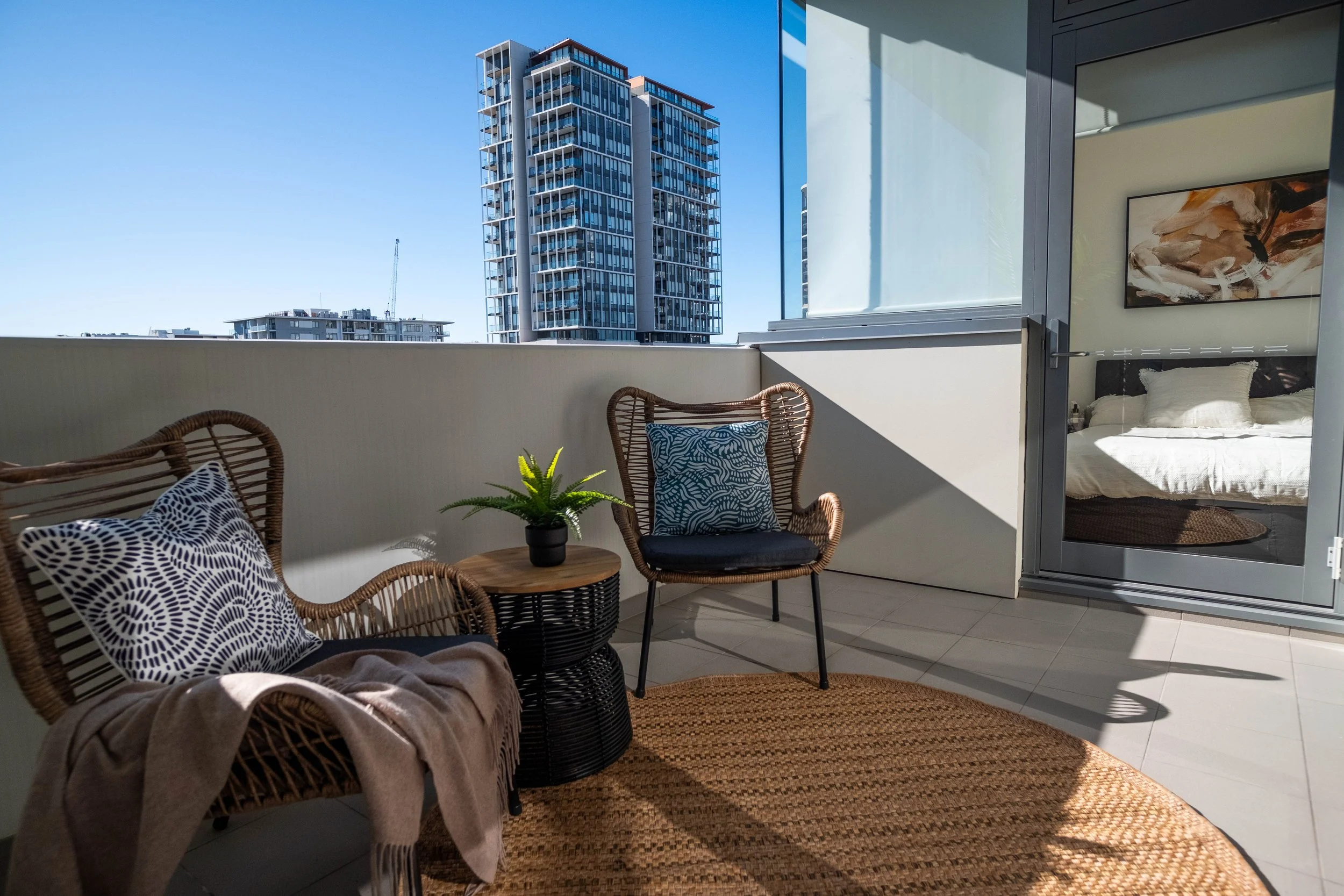 Balcony with wicker chairs, a small round table with a potted plant, and a view of modern high-rise buildings outside. Interior visible includes a bed with white linens and abstract artwork.