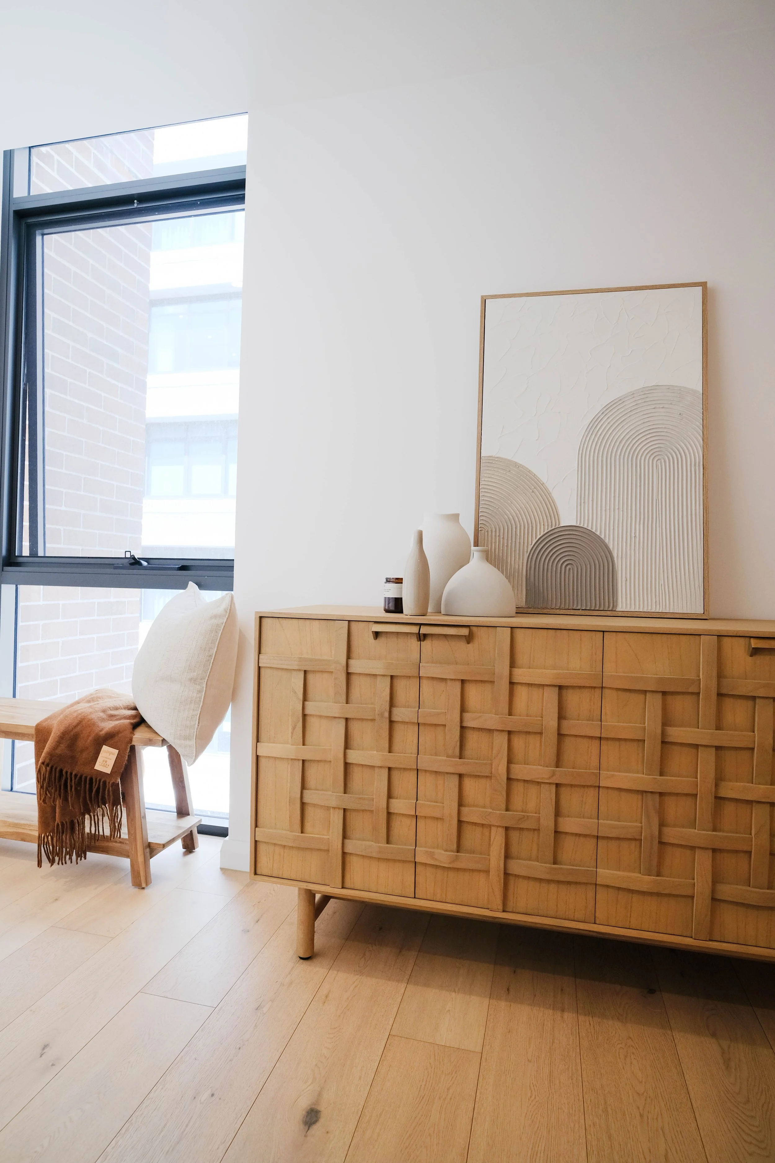 A light wood sideboard with a woven pattern on the doors, topped with several vases and a large framed textured abstract art piece. There is a window with a view of a brick building, and a bench with a cushion and a toss blanket in front of it.