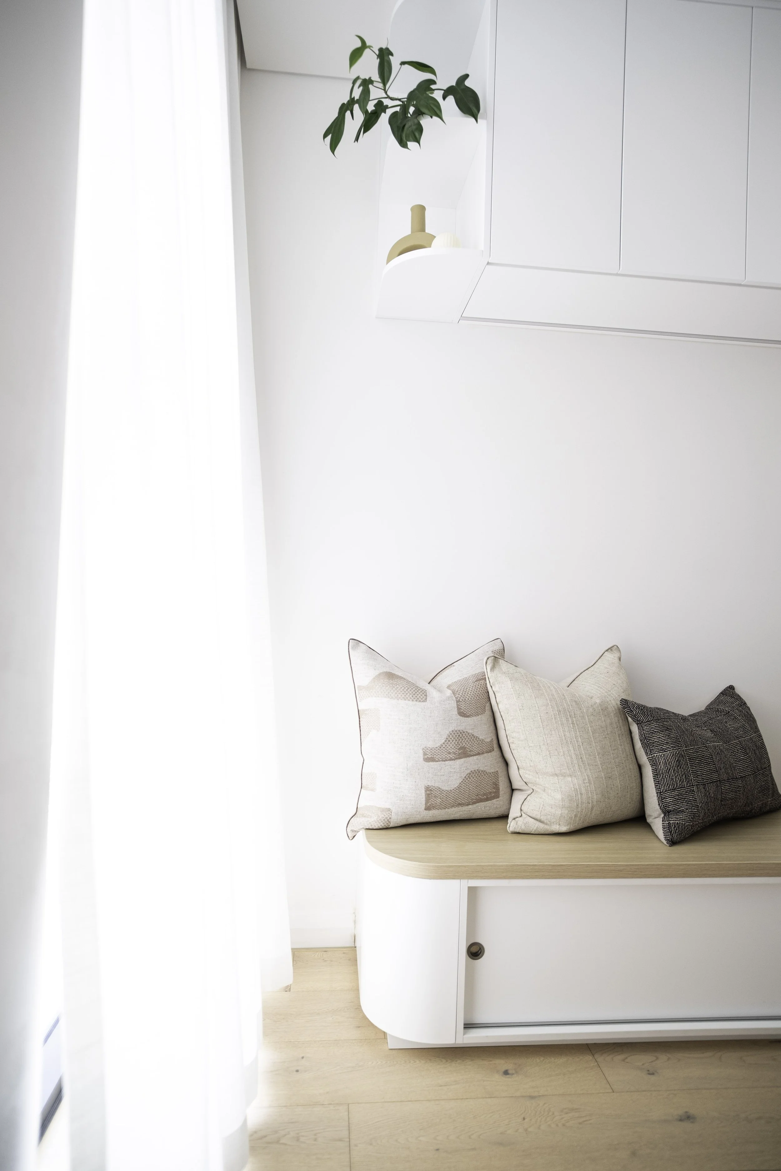 Minimalist white living space with a cozy seating nook decorated with three neutral-colored cushions, natural wood flooring, a sheer white curtain on the left, and a white wall-mounted cabinet with decorative items and a green plant.