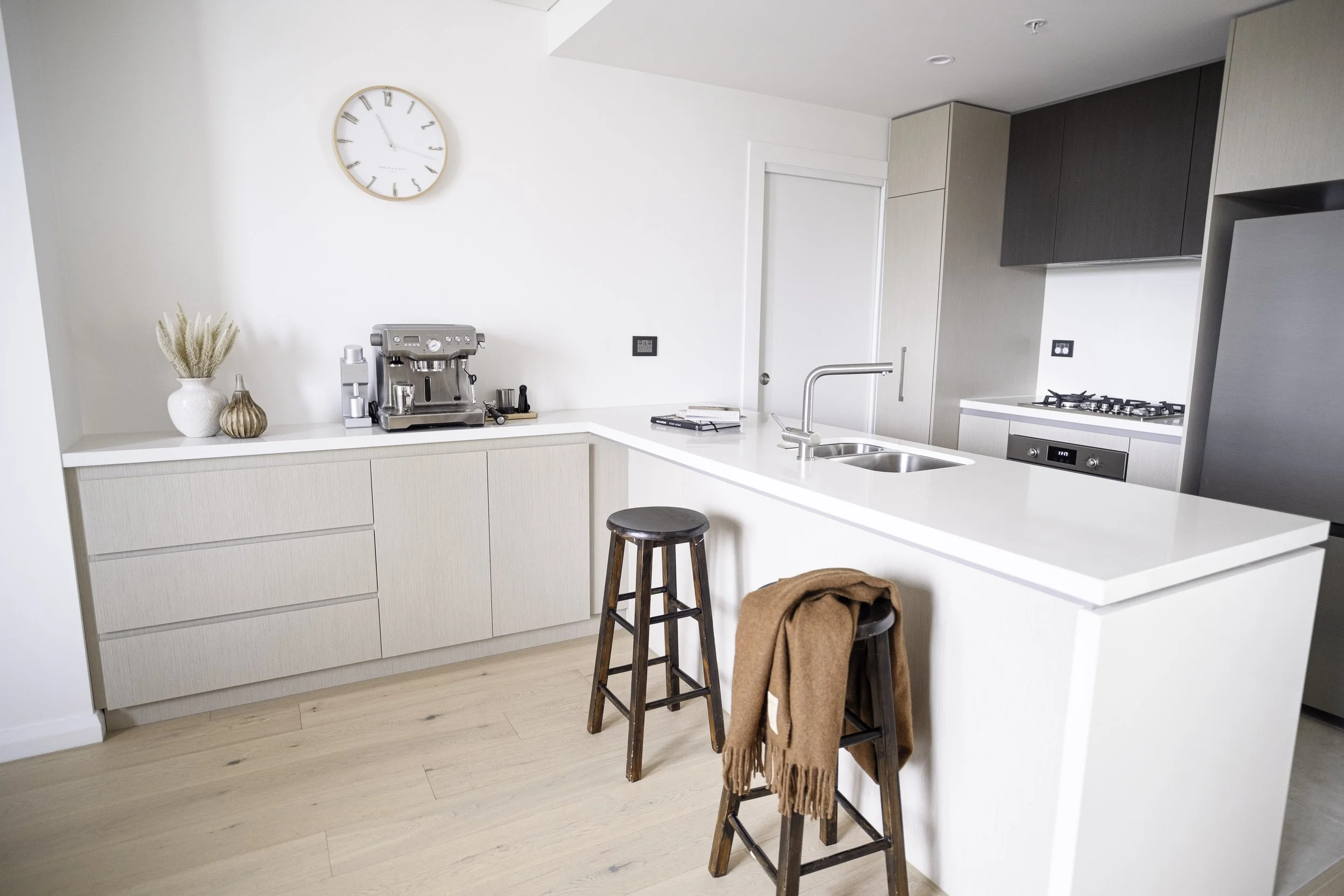 Modern kitchen with white countertops, beige cabinets, and two wooden bar stools, one with a brown scarf. Kitchen appliances include a coffee machine and a stovetop. A wall clock shows 11:03. Decor includes vases with dried plants.