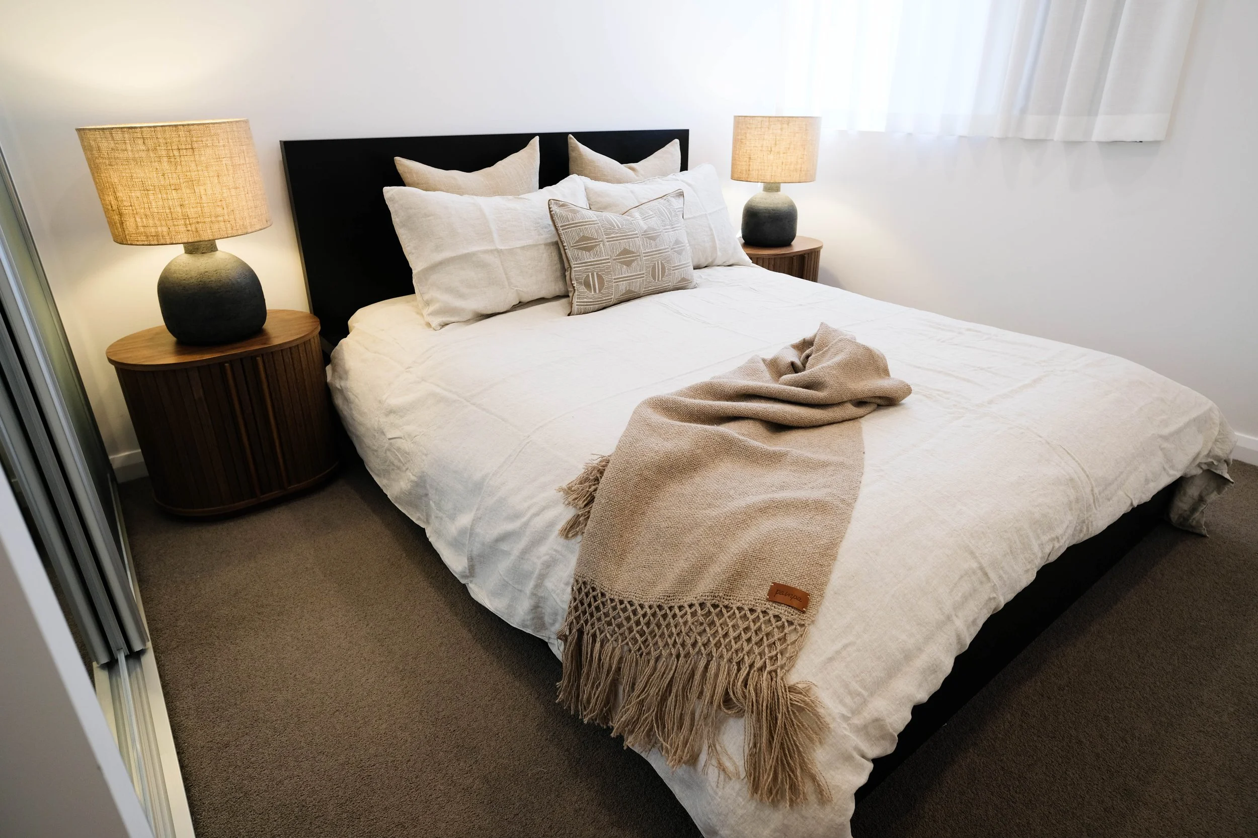 A neatly made bed with a black headboard, white I love Linen bedding, multiple Macey & Moore cushions, a beige Pampa throw blanket, two wooden side table with matching lamps, a window with sheer white curtains.