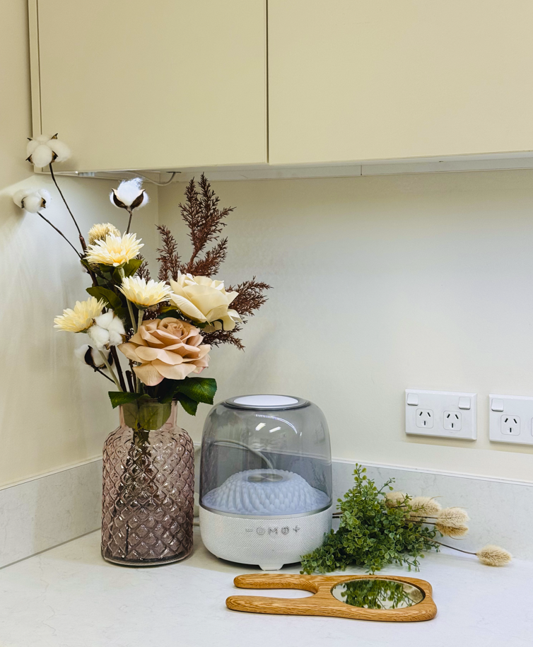 A kitchen countertop with a pink textured vase holding artificial flowers, a white diffuser with a glass cover, a small green leafy sprig, and a wooden hand mirror.