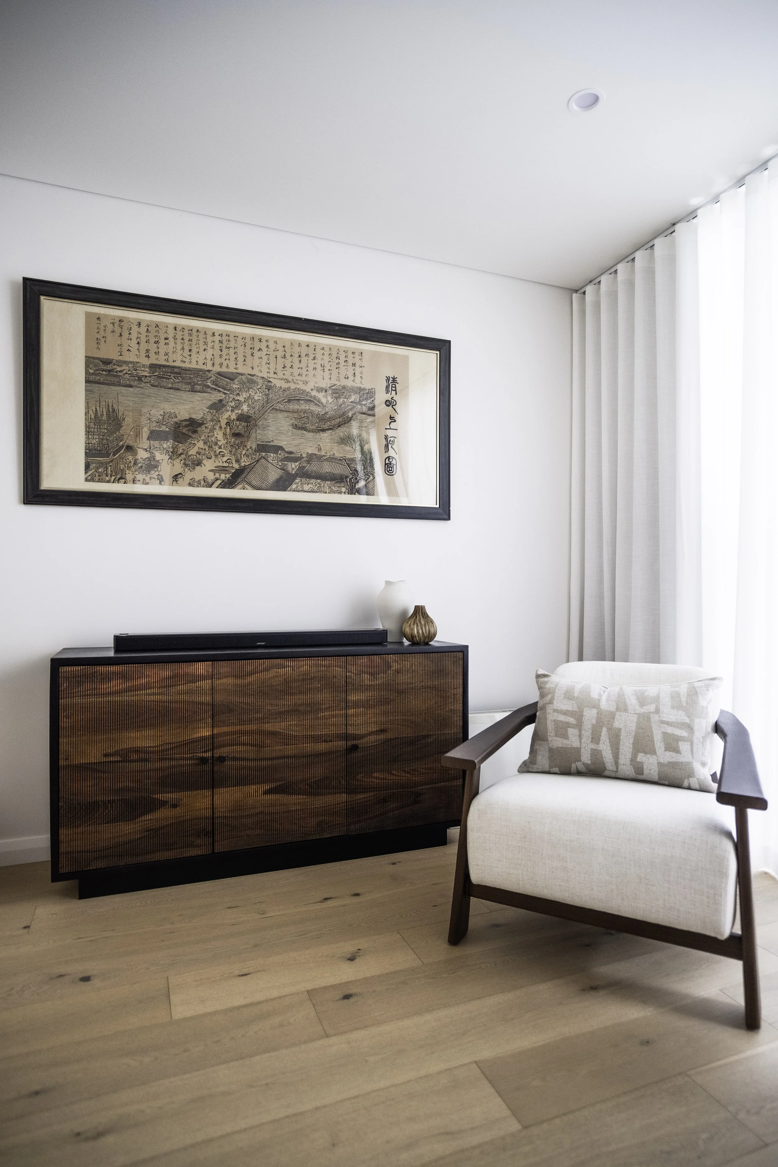 A modern living room corner with a framed Asian art print hanging above a Horgans wooden sideboard, a cream Horgans armchair with a Macey & Moore patterned pillow, and a sheer white curtain covering a window in the background.