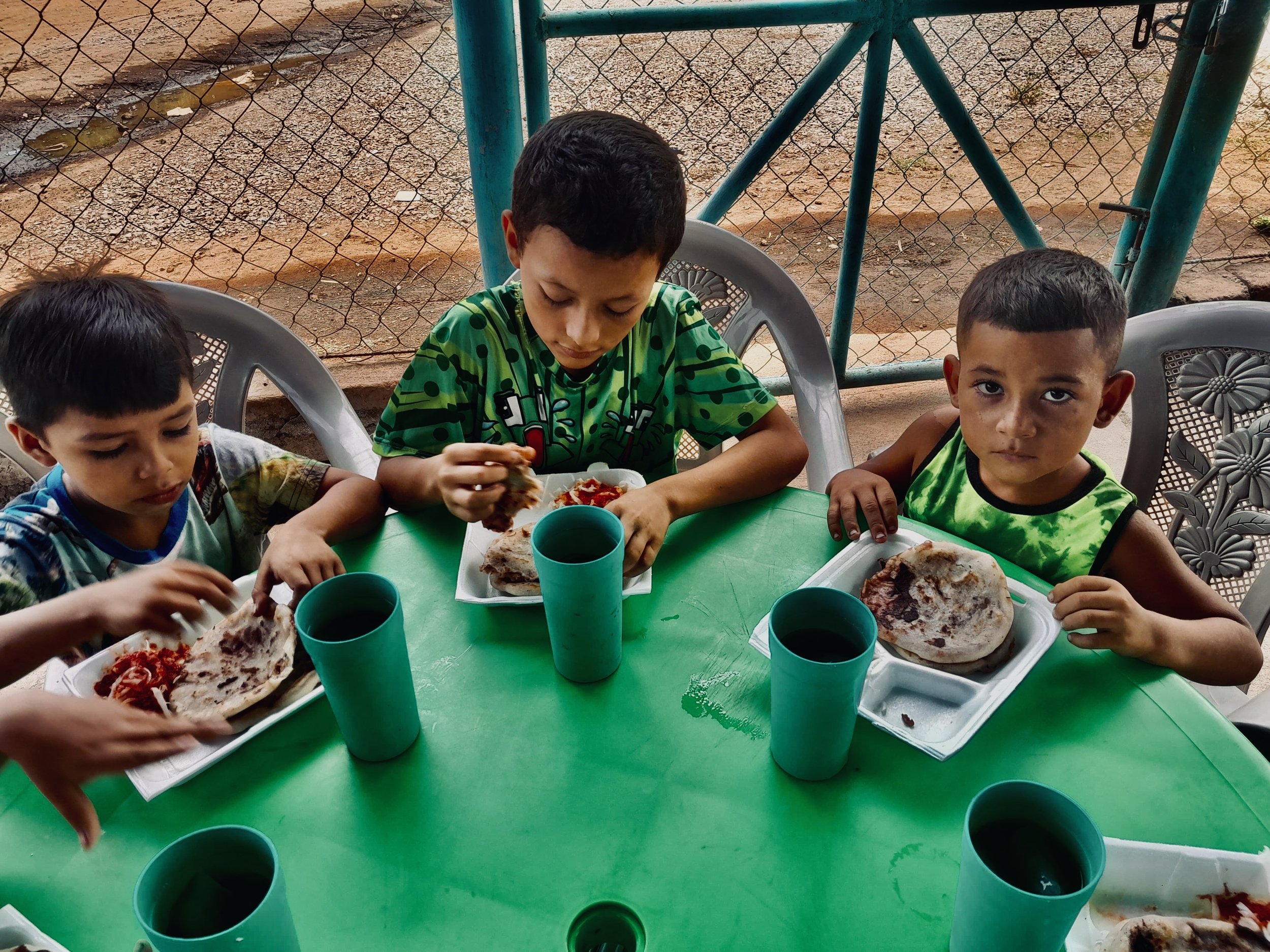 Three children sitting at a green table, eating pizza and drinking dark-colored beverages in cups.