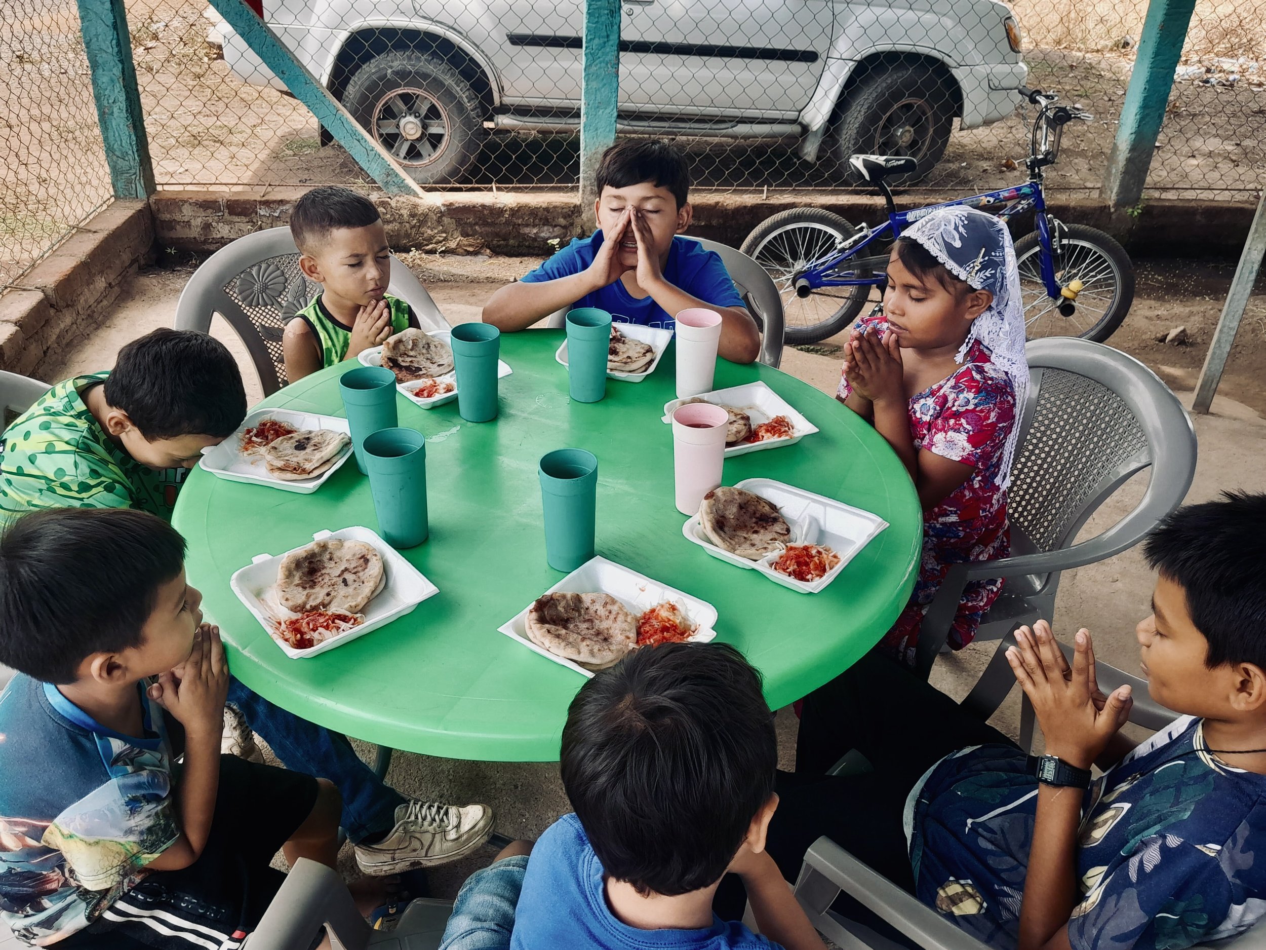 Children praying around a green table with plates of food and cups, outdoors in a fenced area.