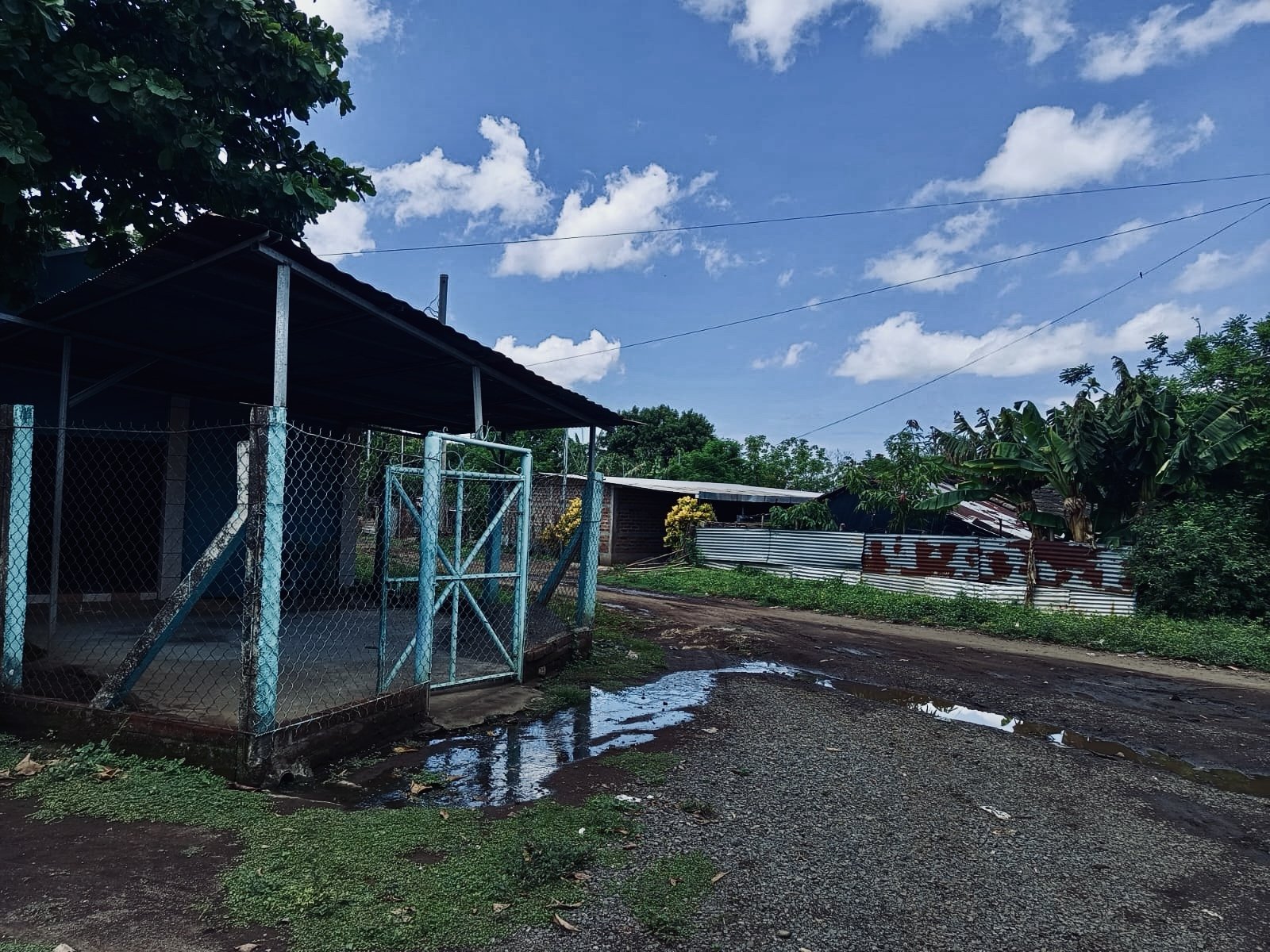 A dirt road with puddles, a chain-link fence with a gate, a small covered structure, lush green trees, and houses with corrugated metal roofs under a partly cloudy sky.
