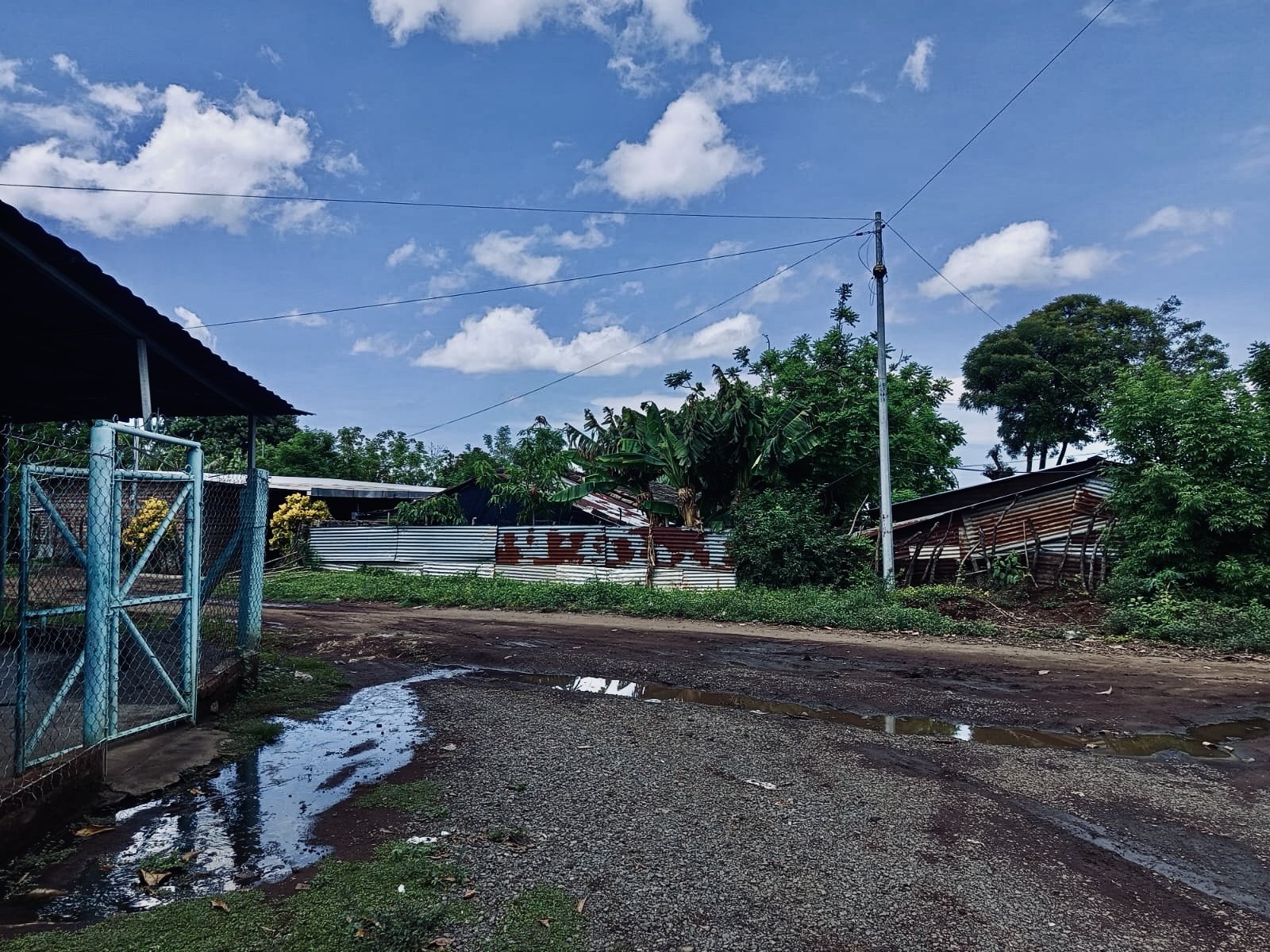 A dirt and gravel road with puddles, surrounded by trees and simple houses with corrugated metal roofs. A utility pole with wires, a blue gate on the left, and a partly cloudy sky overhead.