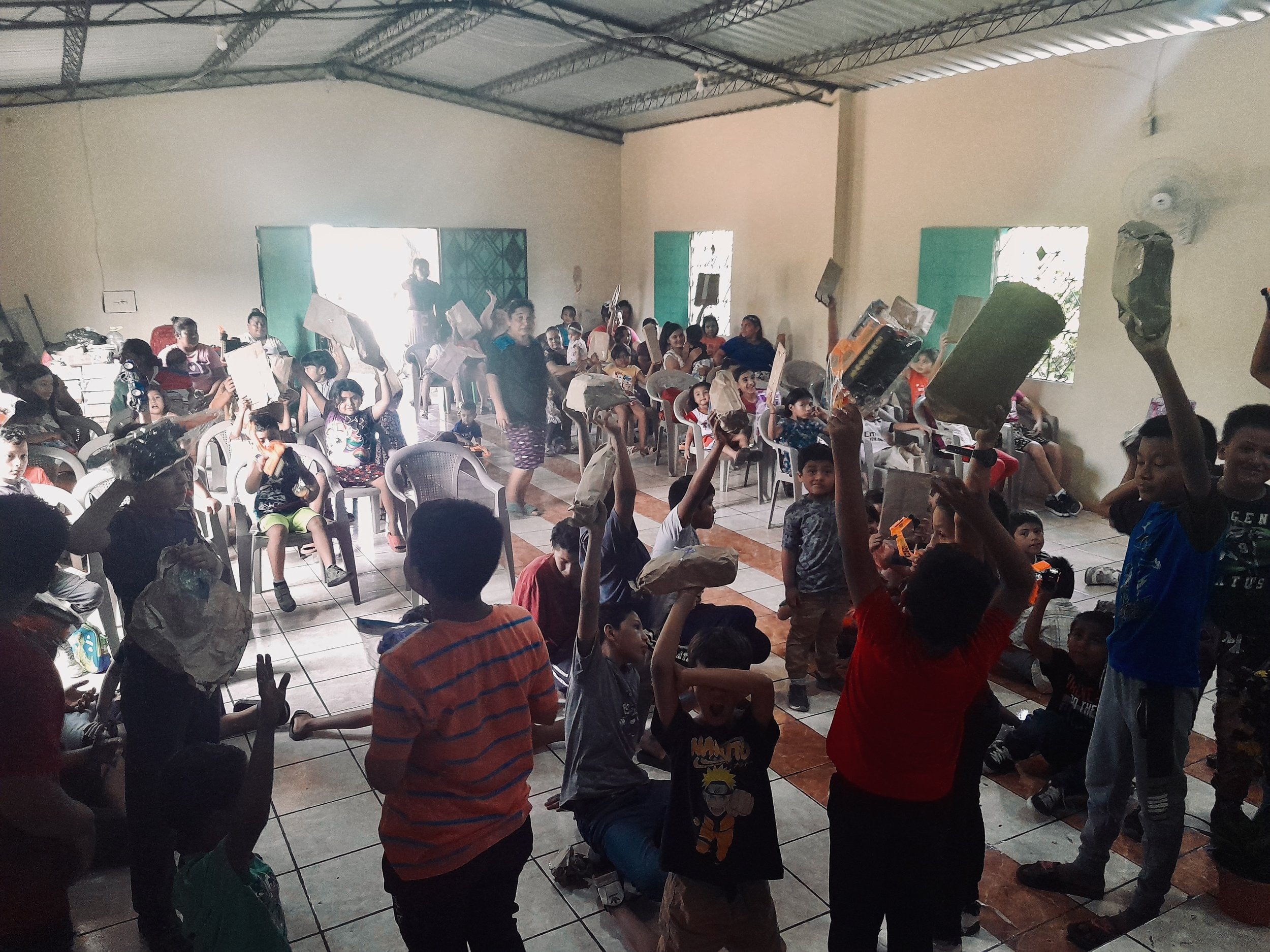 Children and adults gathering indoors, some seated and others standing, holding up items like toys and books, with bright light coming from an open door in the background.