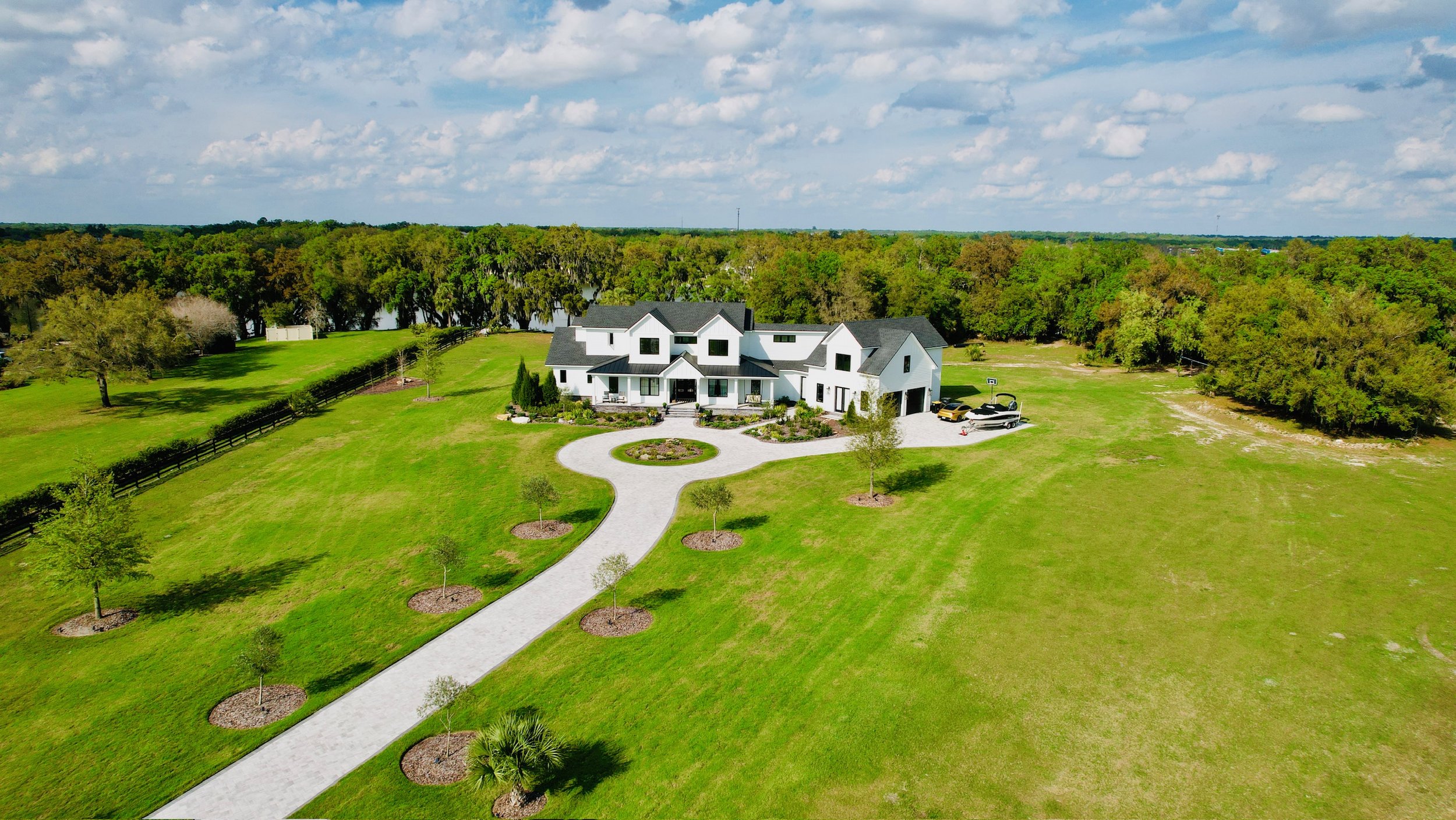 A large white modern house surrounded by green lawns and trees, with a circular driveway and a small parking area with vehicles, set against a backdrop of a lake and a partly cloudy sky.