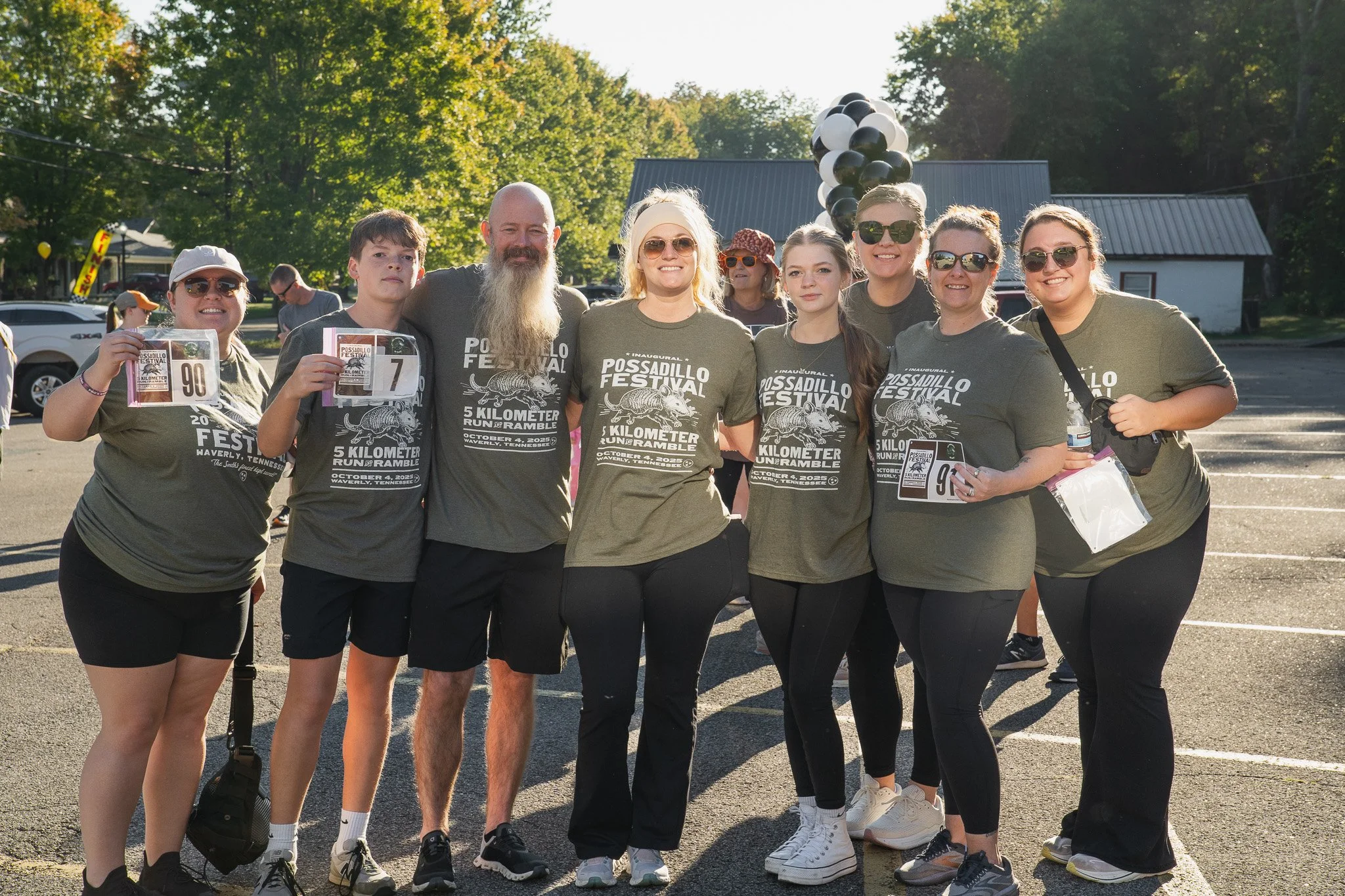 Group of people in matching t-shirts at the Possadillo Festival 5 Kilometer Run in Waverly, Tennessee, posing outdoors with trees and balloons in the background.