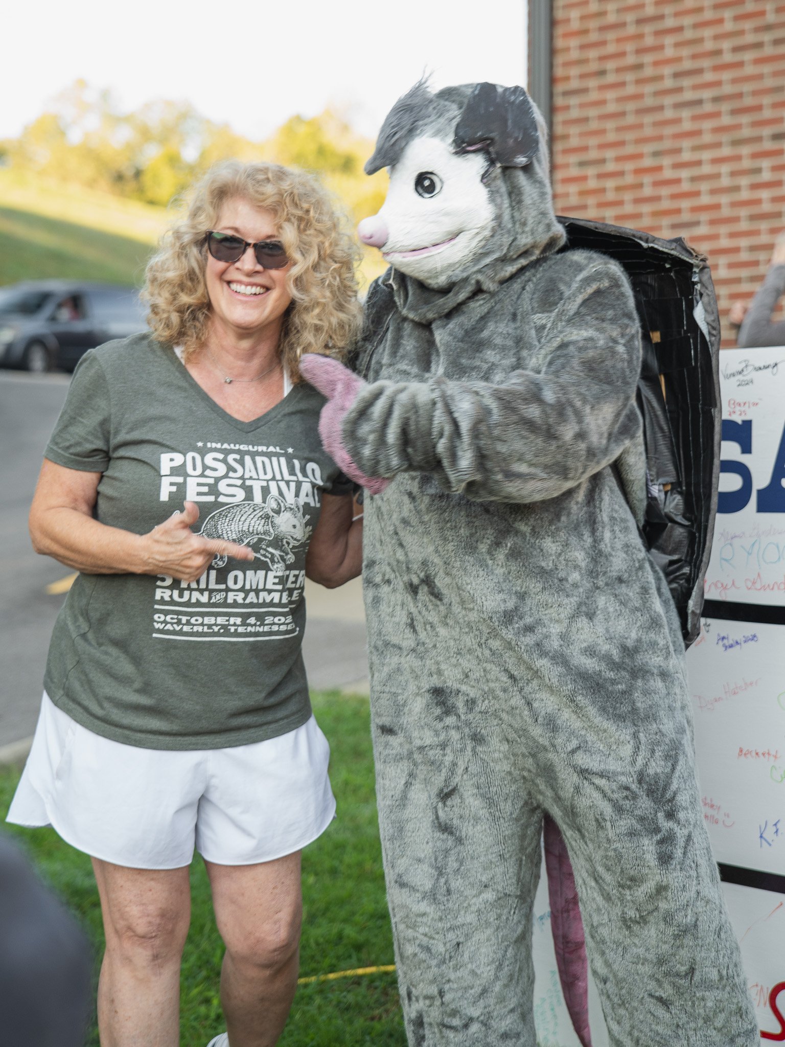 Woman with curly hair and sunglasses pointing to a person dressed in a large animal costume.