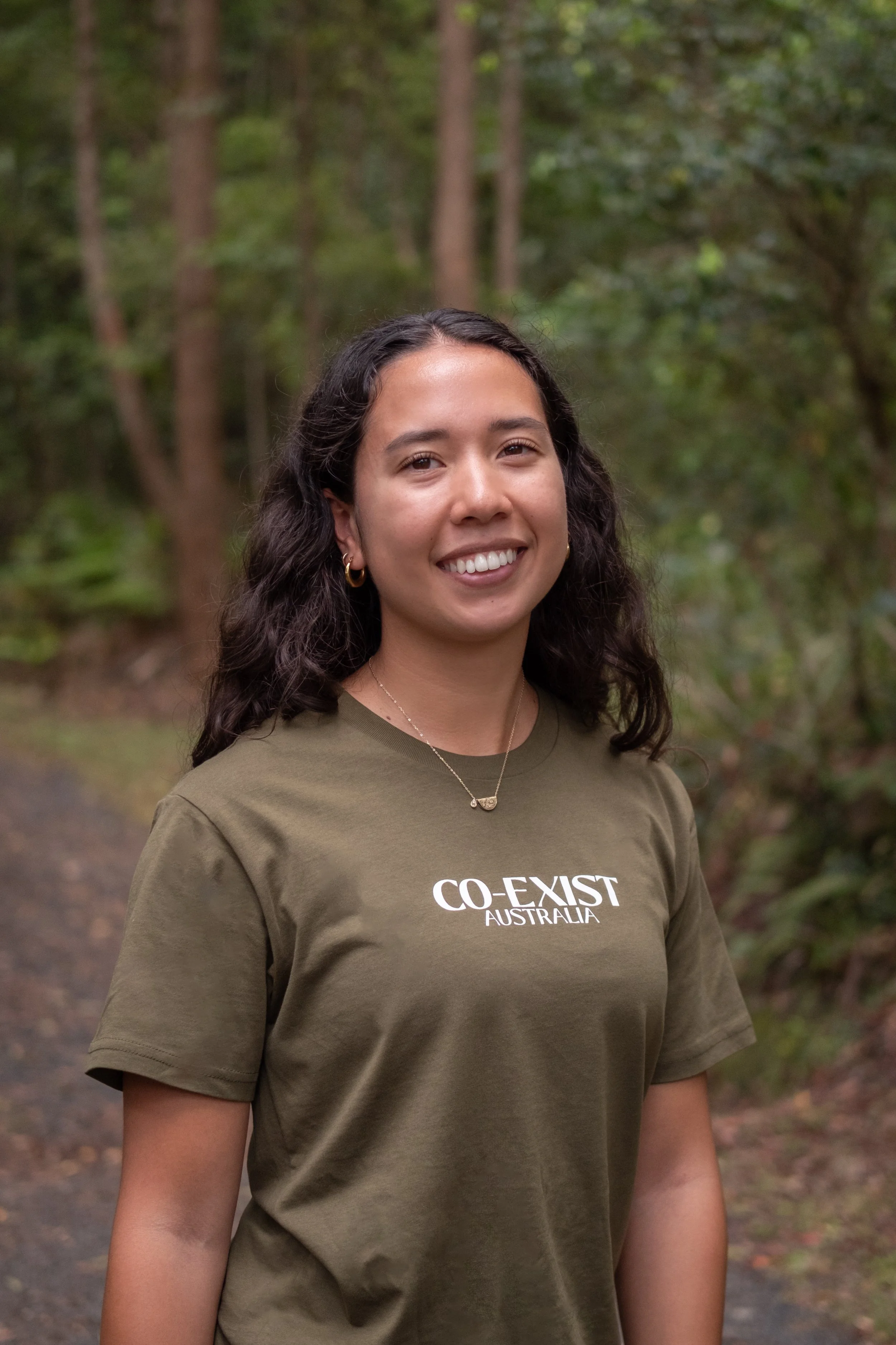 Smiling person in front of green foliage, wearing a necklace and earrings.