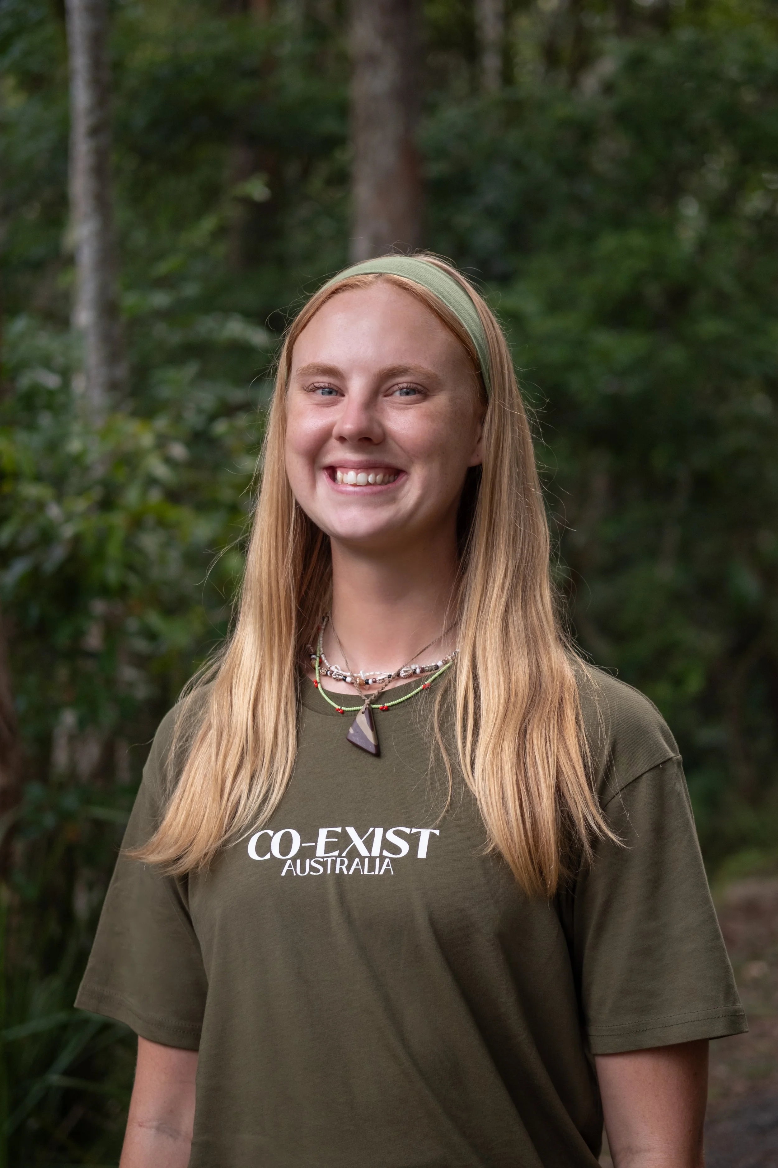 Young woman with long blonde hair standing in front of green foliage, wearing a necklace and earrings, smiling slightly.