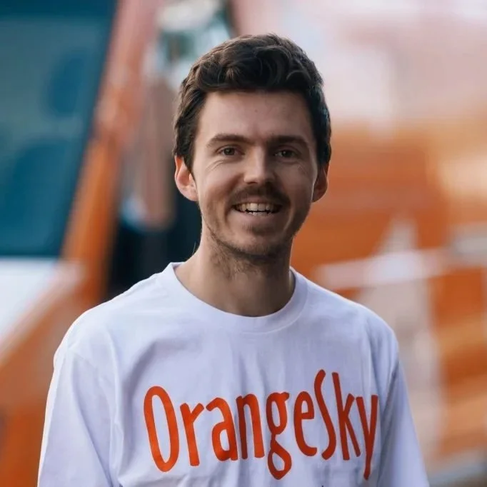 Man wearing a white Orange Sky Australia t-shirt, smiling, with an orange vehicle blurred in the background.