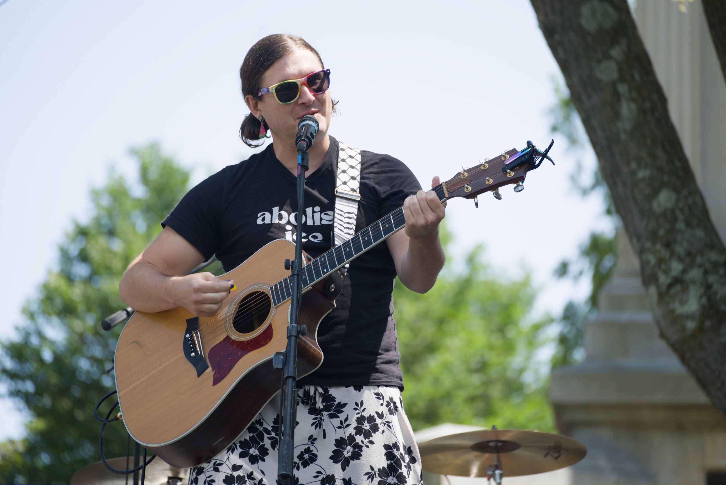 A white woman with brown hair wearing sunglasses and a black shirt that says “Abolish ICE” holding a guitar and standing behind a microphone.