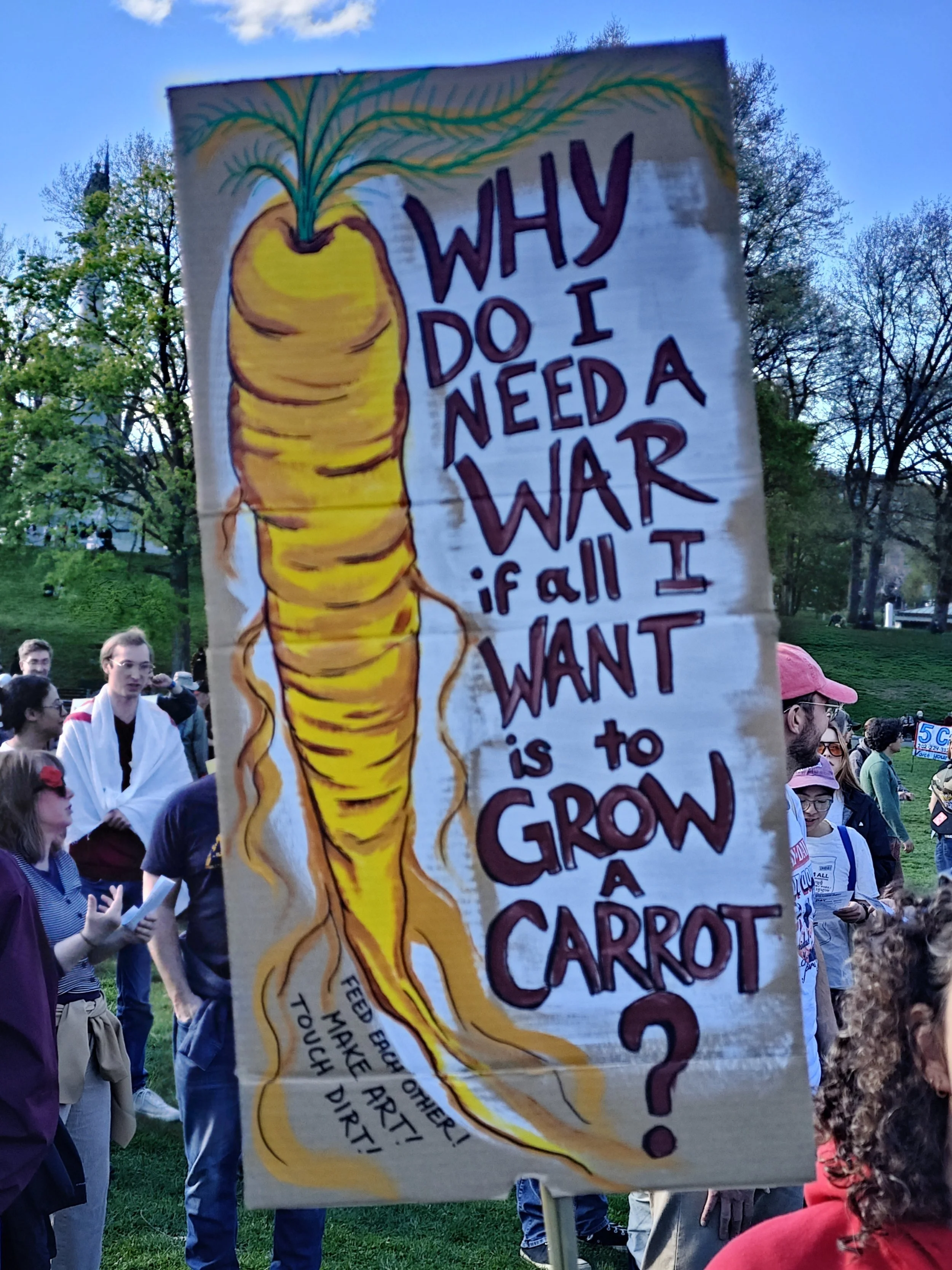 A large protest sign with a beautifully hand painted carrot that says "Why Do I Need A War if all I WANT is to GROW a CARROT? Feed each other! Make Art! Touch Dirt!"