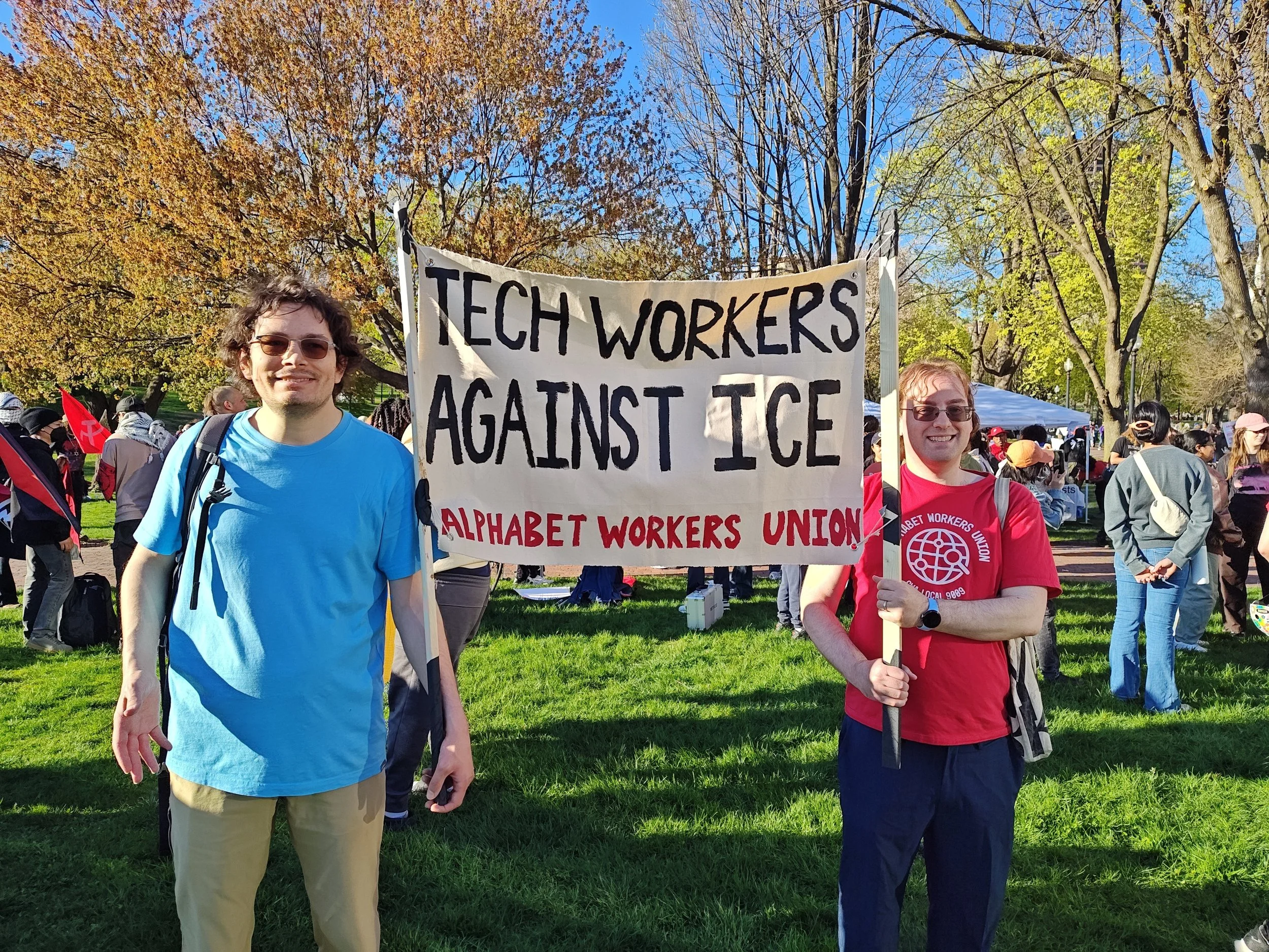 Two smiling protestors holding a banner saying "Tech Workers Against ICE: Alphabet Workers Union"