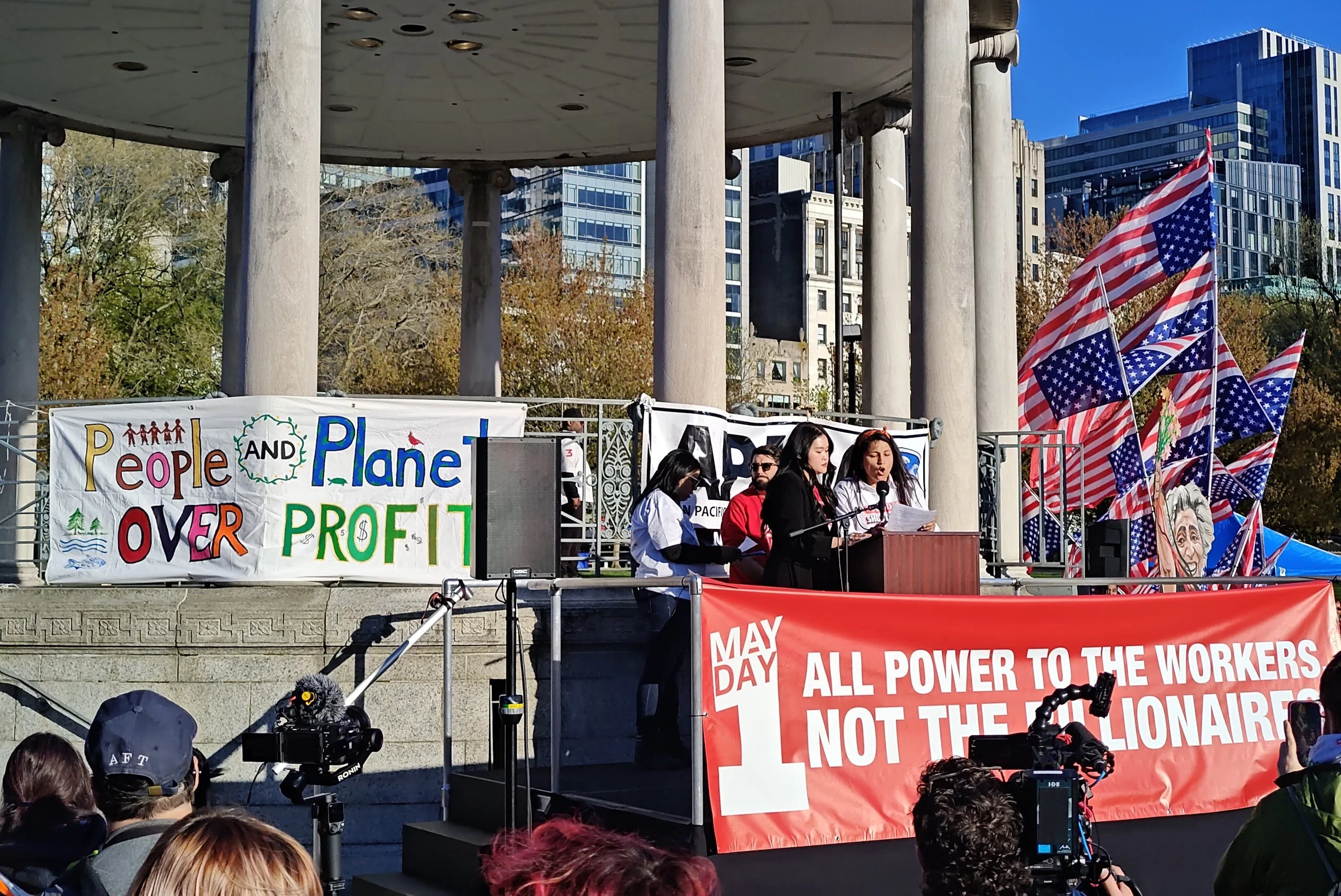 People speaking at the Parkman Bandstand with signs saying “People and Planet over Profit” and “May Day: All power to the workers, not the billionaires”.