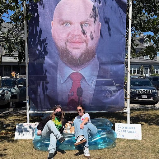Outside, two people sit smiling on an inflatable couch with a stuffed cat in front of an 8x10 foot image of the bald JD Vance meme.