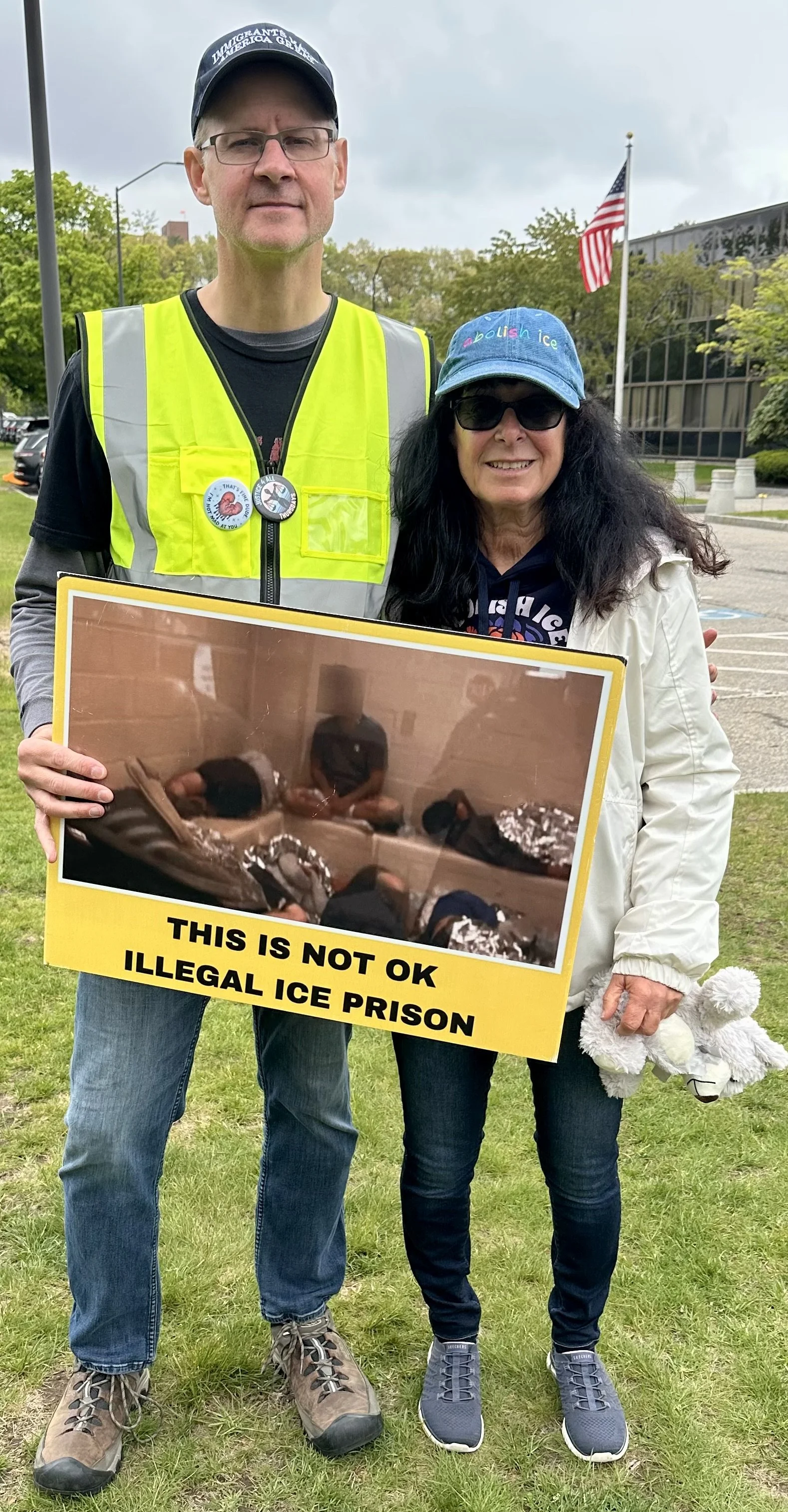 A tall white man standing next to a white woman and holding a sign saying “This is not OK. Illegal ICE Prison” beneath a photograph of men sitting and laying in a window-less room.