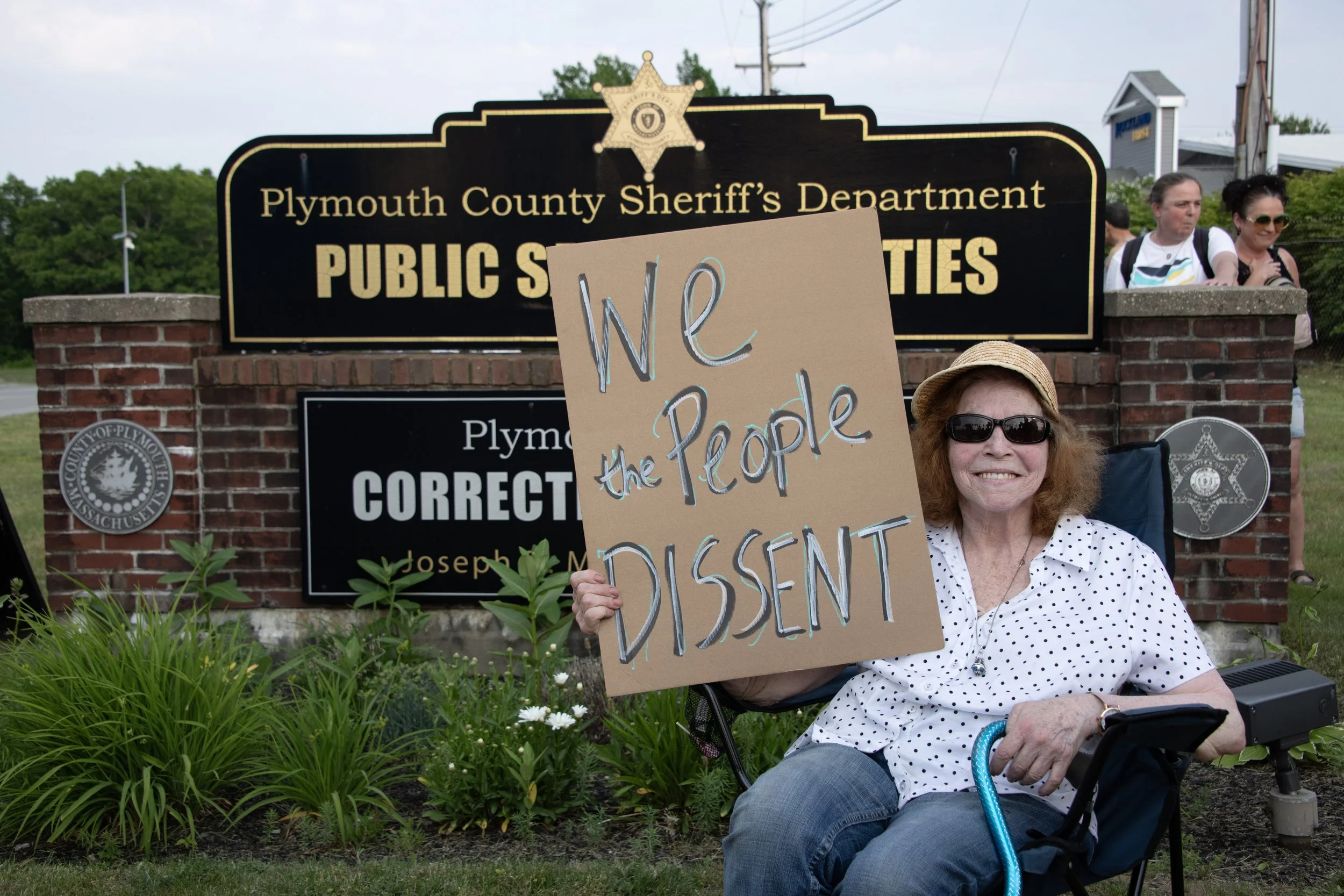 A woman holding a sign saying “We the People Dissent” in front of the Plymouth County Correctional Facility.