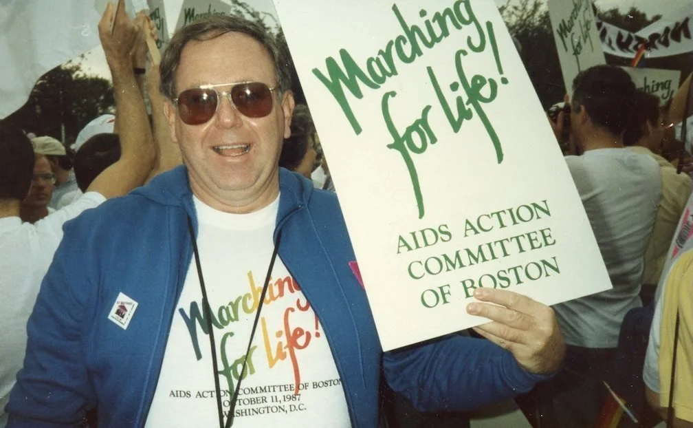 A white man wearing sunglasses, a blue jacket, and a white t-shirt with “Marching for Life!” in rainbow-colored text holding a sign with “Marching for life! AIDS Action Committee of Boston” printed in green ink.