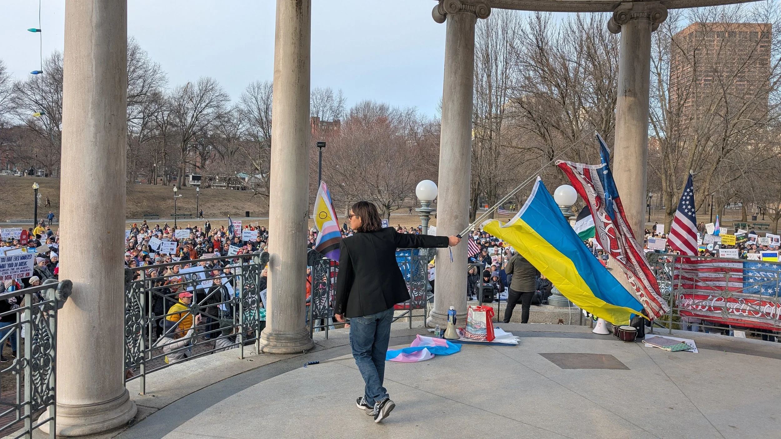 Member of 50501 holding a flagpole with both the American and Ukranian flags on the bandstand in the Common. There is also a progress pride and trans pride flag in the background.