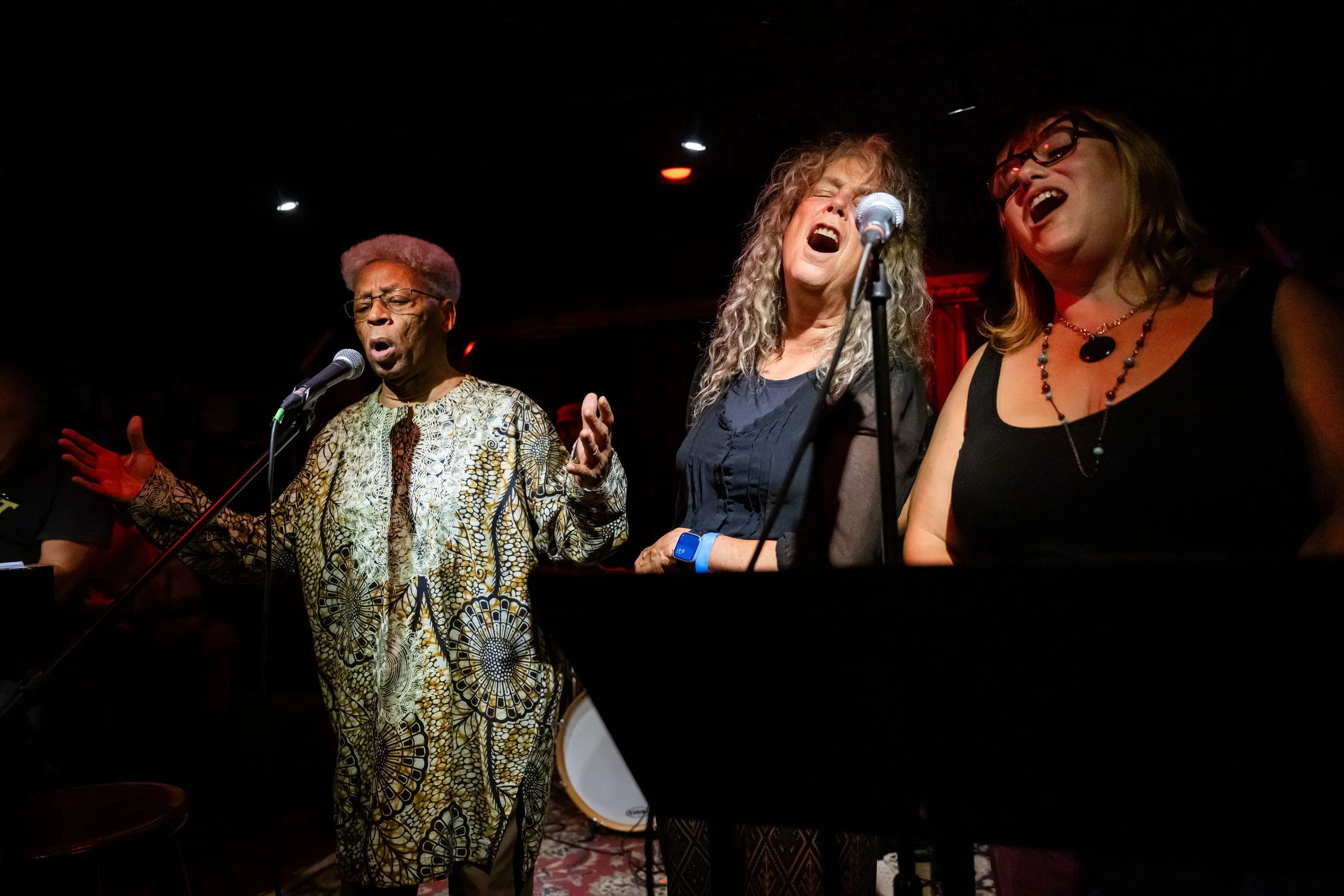 Three people singing on a stage with a dark background, performing as part of the concert series Which Side?: A Protest Music Teach-Out.