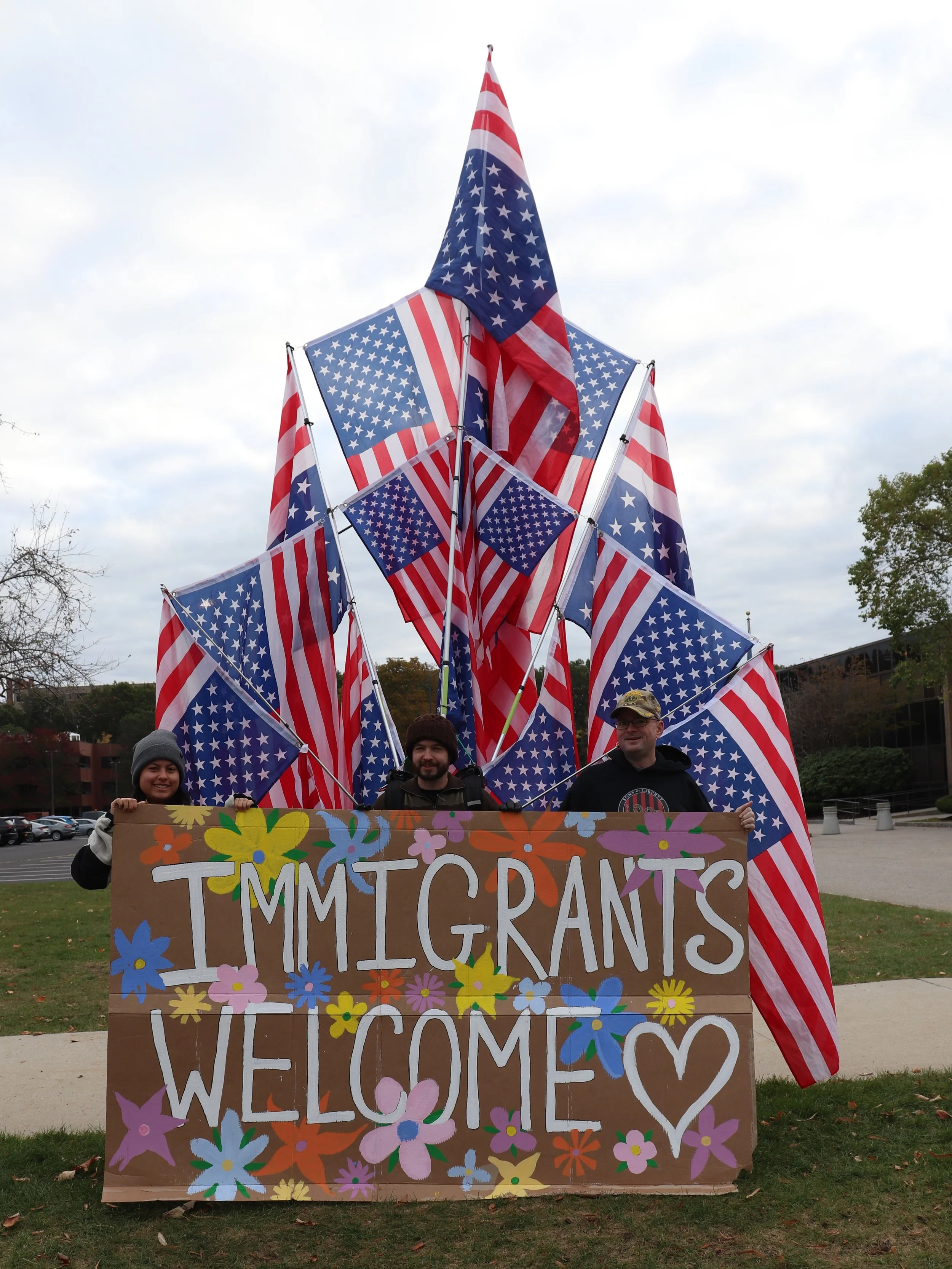 Set of many american flags flying upside down behind a sign that says "Immigrants Welcome" held by MA50501 volunteers