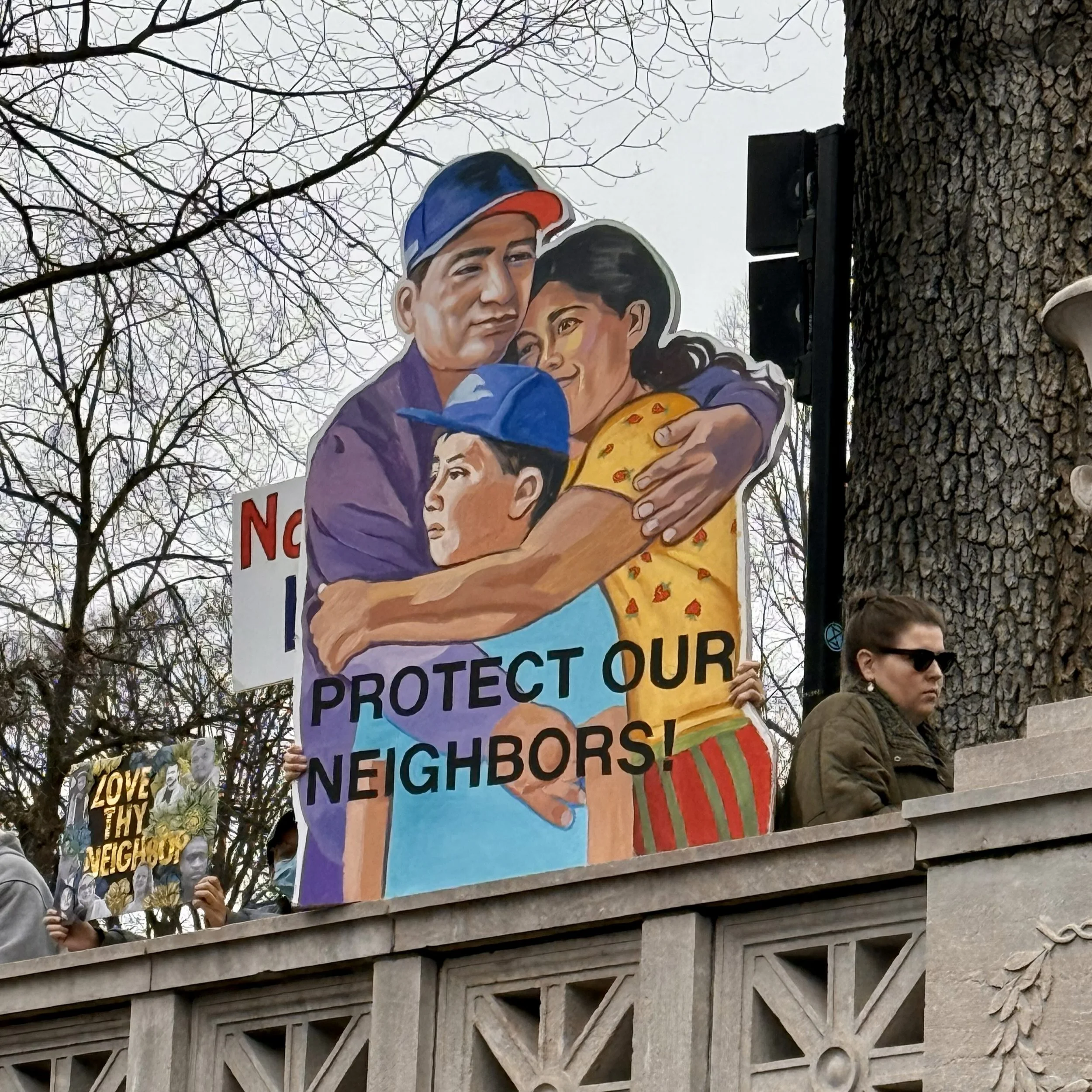 A painted sign at a protest depicting a latino family hugging saying "Protect our neighbors"
