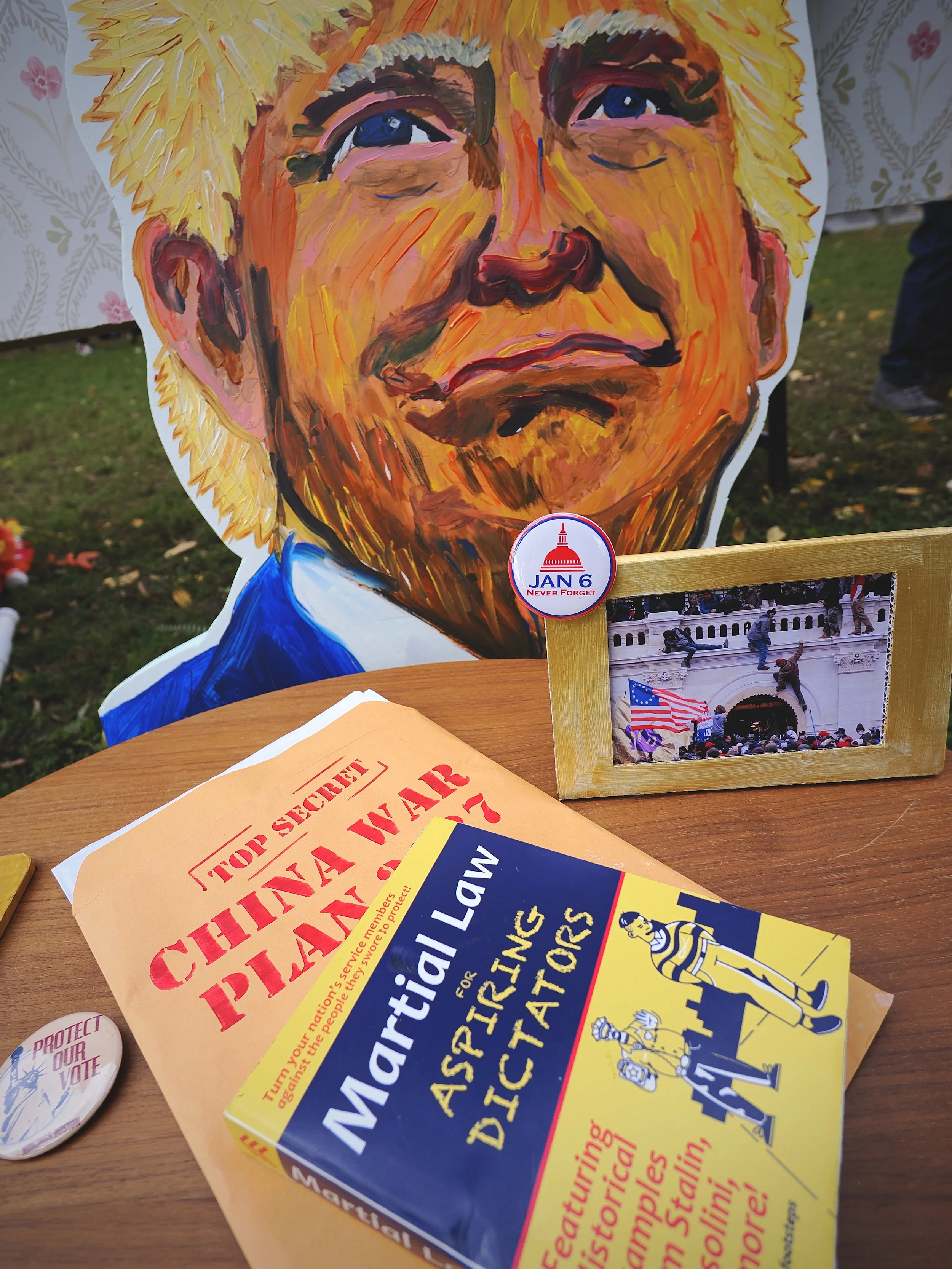 A painting of Donald Trump behind a photograph of the January 6th insurrection, a folder labeled “Top Secret: China War Plans”, and a book titled “Martial Law for Aspiring Dictators.”