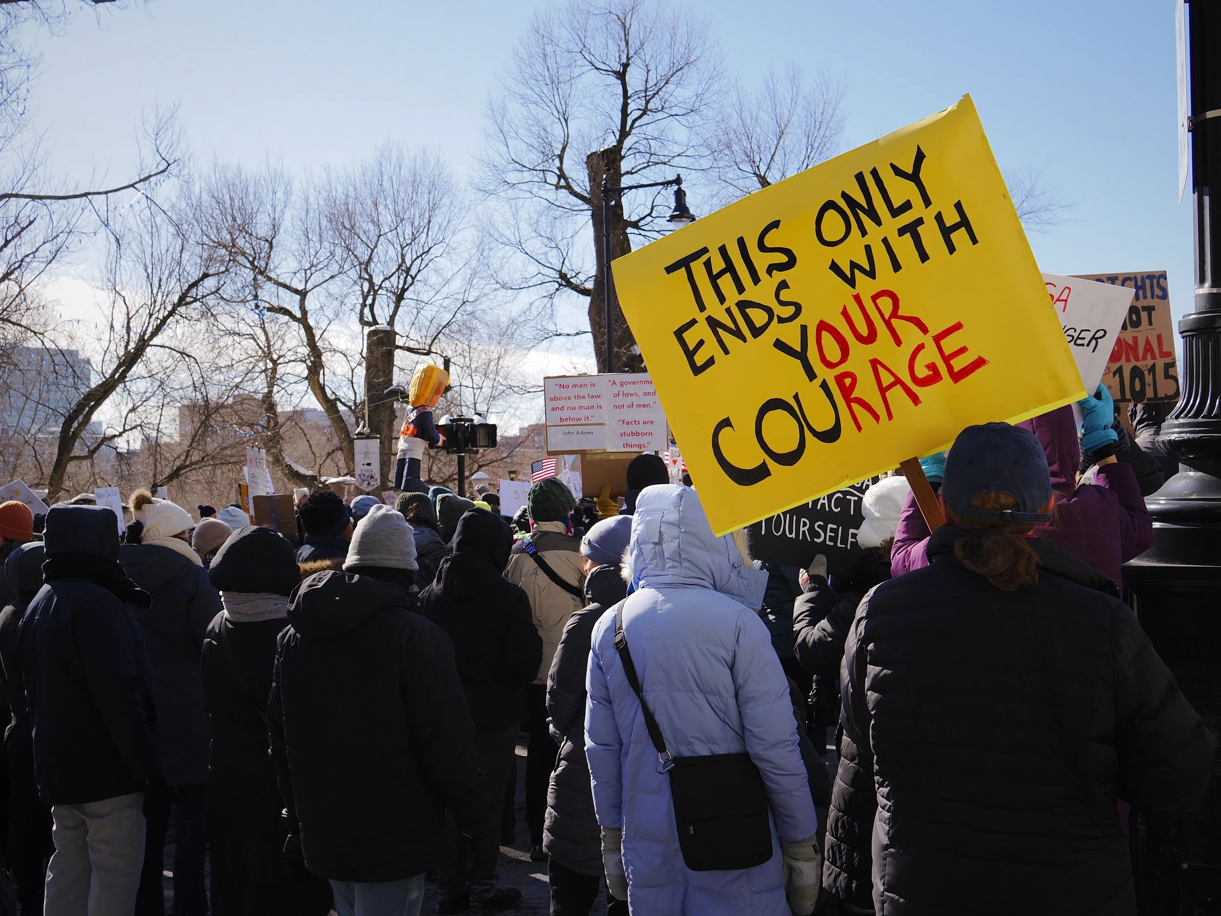 A Mass 50501 community gathering of Massachusetts residents showing a bright yellow sign that says "This Only Ends With Your Courage" and the letters spelling "Our Rage" painted red.