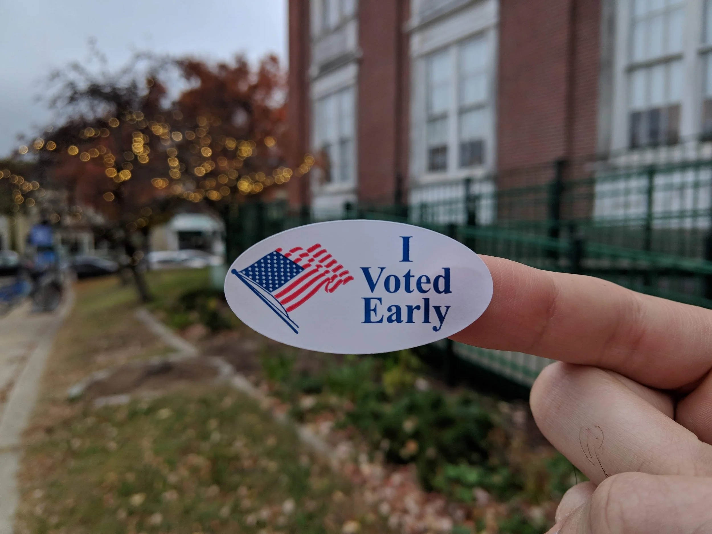 A sticker with text saying “I voted early” held in front of some autumnal trees and an early-voting polling place.