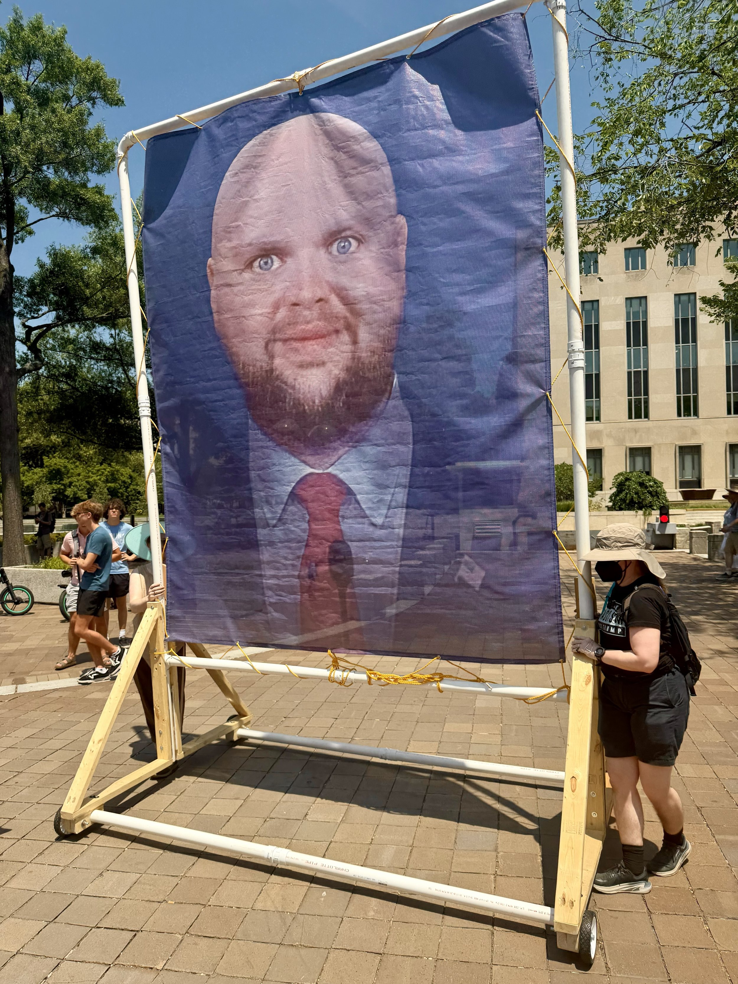 A large banner with a satirical version of J.D. Vance’s head, modified to have his head shaved and a vacant, unintelligent expression on his face, on a frame pushed by two volunteers.