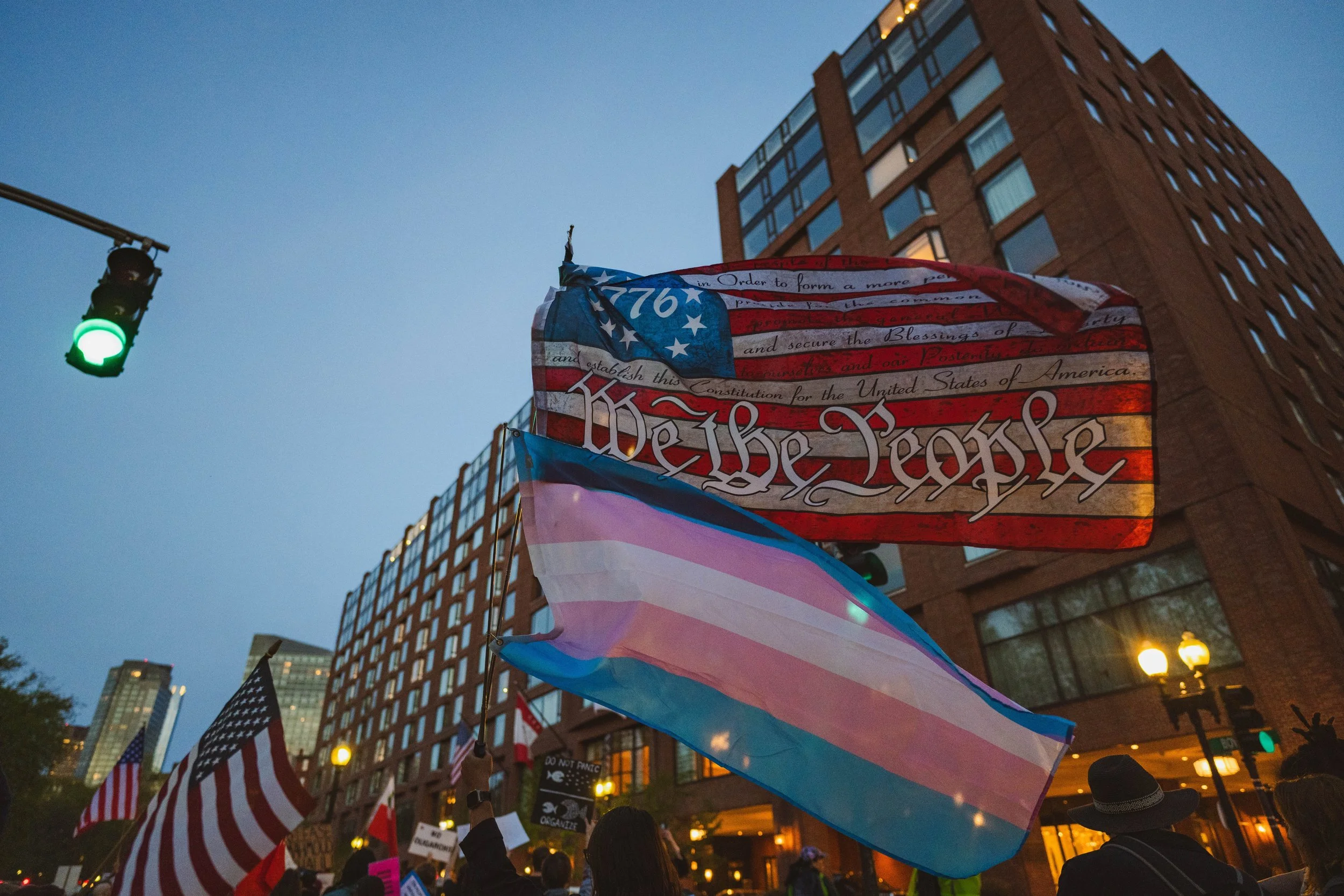 Protestors marching the streets of Boston with a "We the People" and trans rights flag at twilight