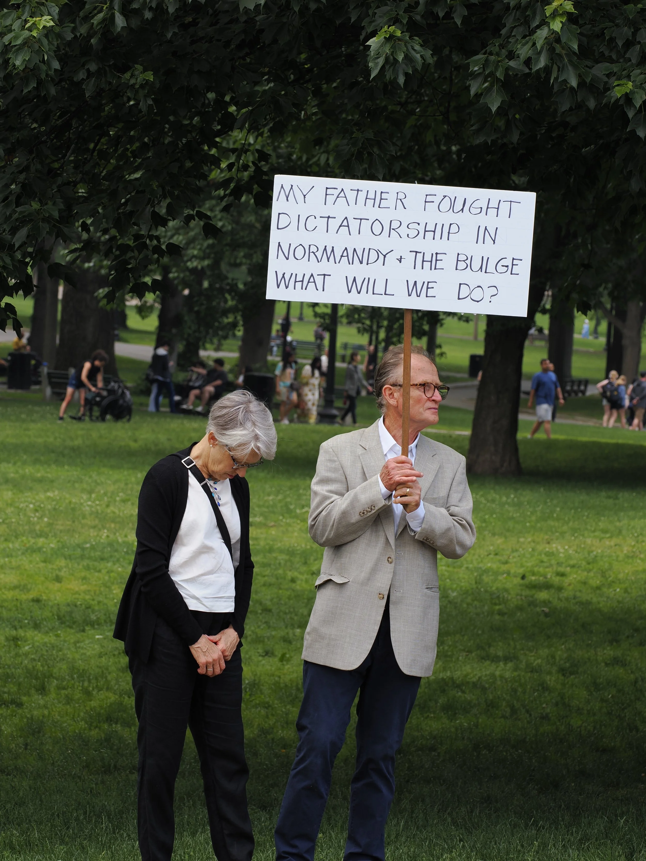 A couple holding a sign at a protest saying "My father fought dictatorship in Normandy + the Bulge. What will we do?"