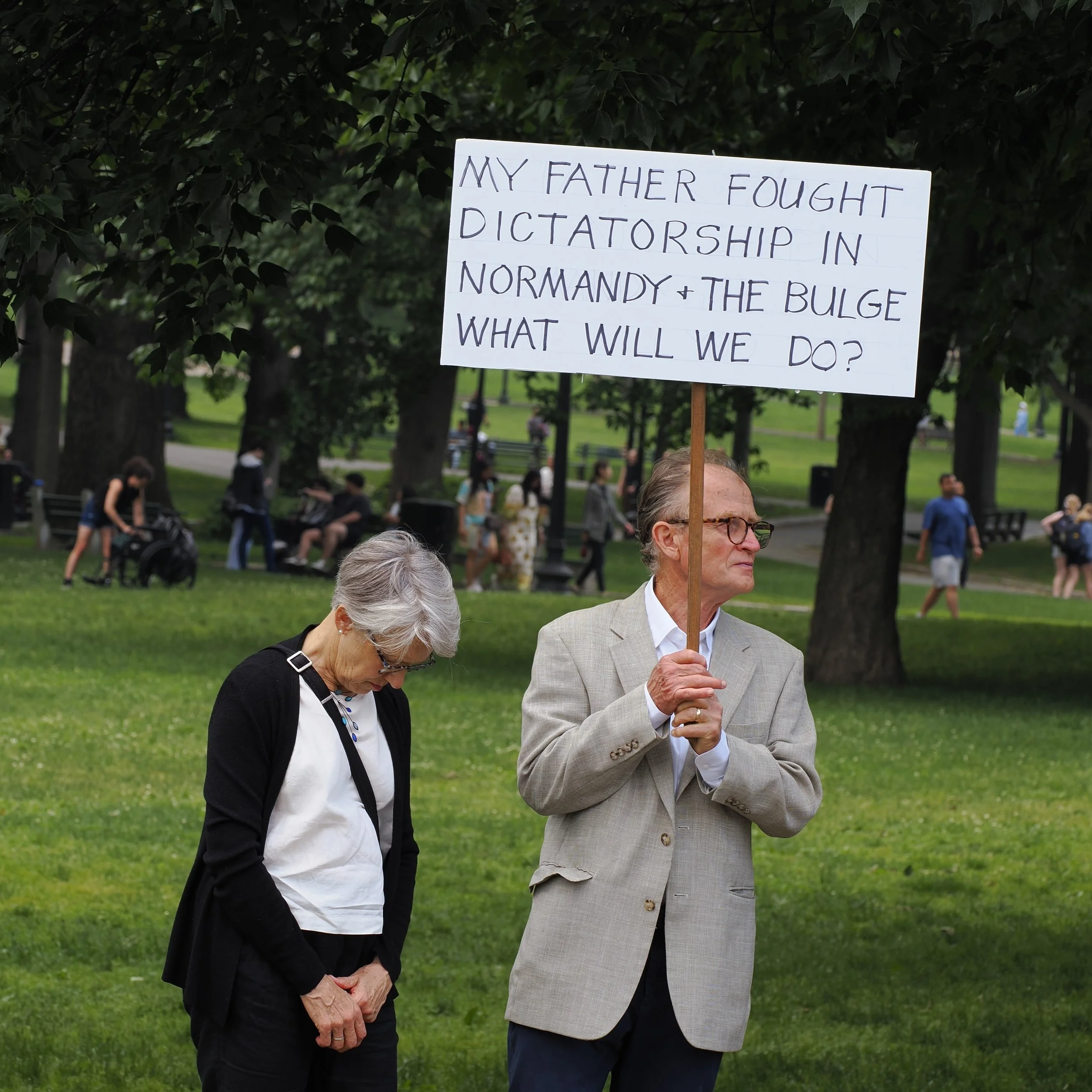 Older man holding sign saying "My father fought dictatorship in Normandy + The Bulge. What will we do?"