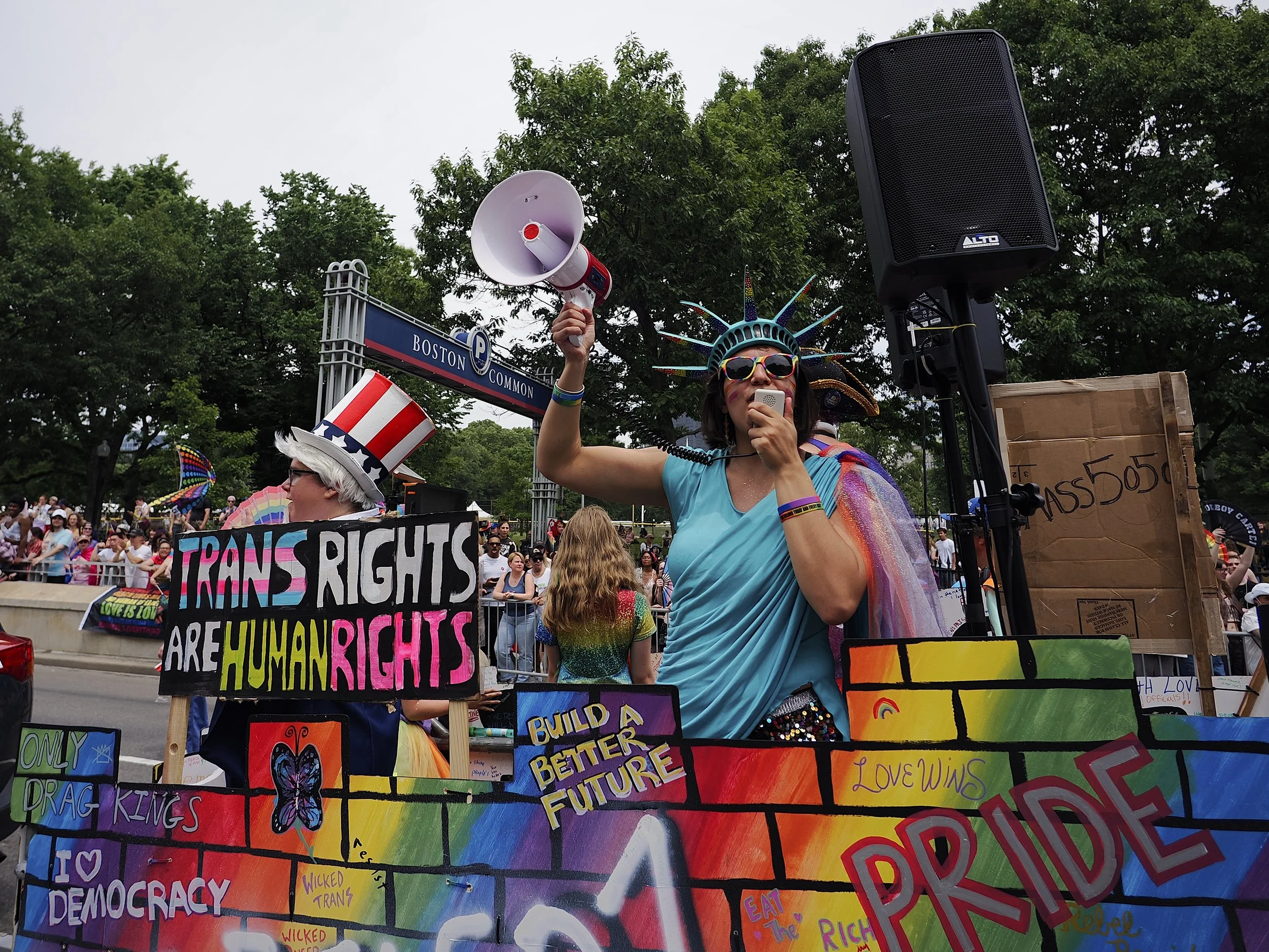 A woman dressed as the statue of liberty shouting into a microphone standing next to a drag king Uncle Sam on a rainbow-colored float painted with slogans such as “trans rights are human rights”, “build a better future”, and “only drag kings”.