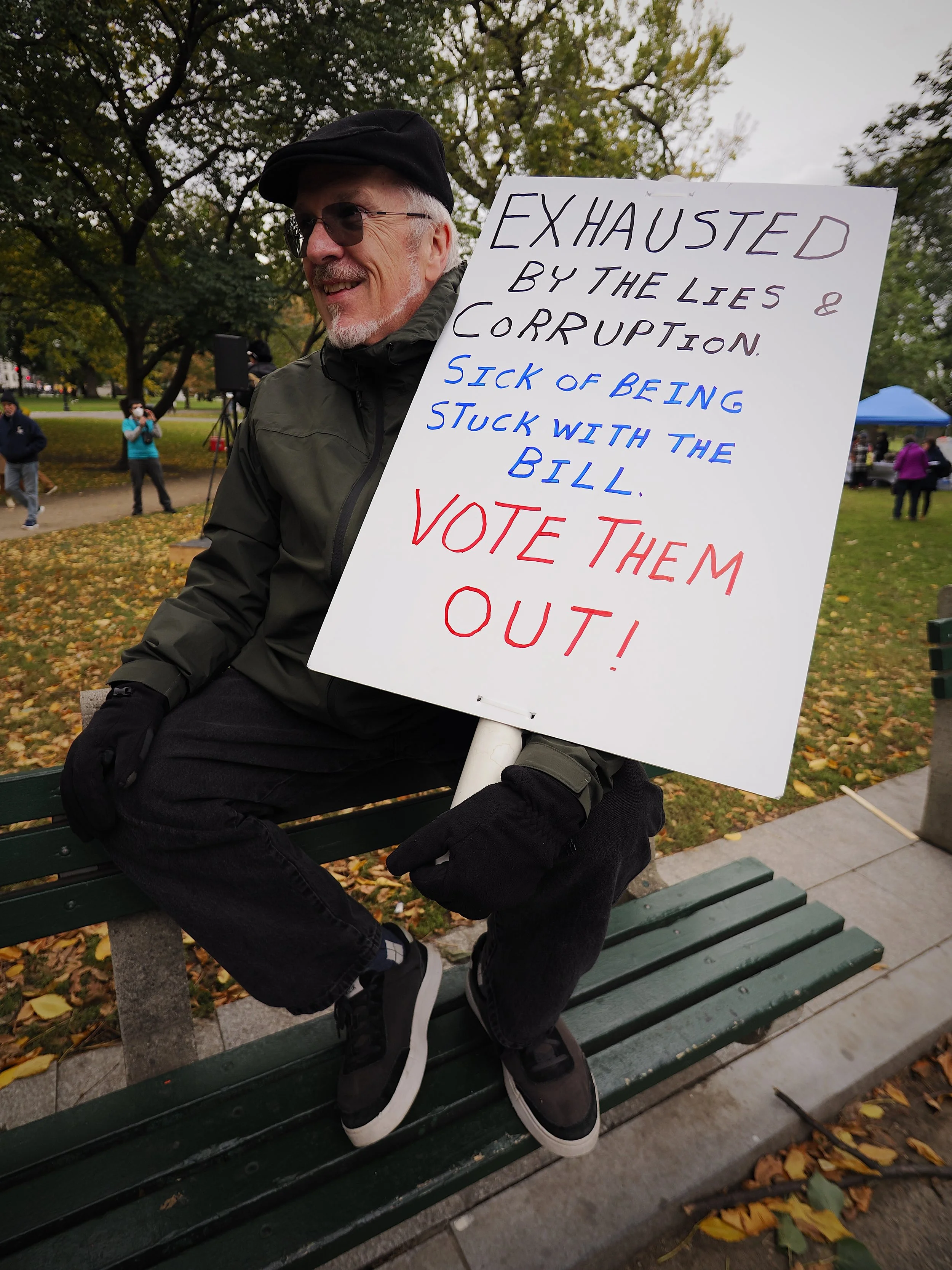 A man sitting on a bench, holding a sign saying, “Exhausted by the lies & corruption. Sick of being stuck with the bill. Vote them out!”