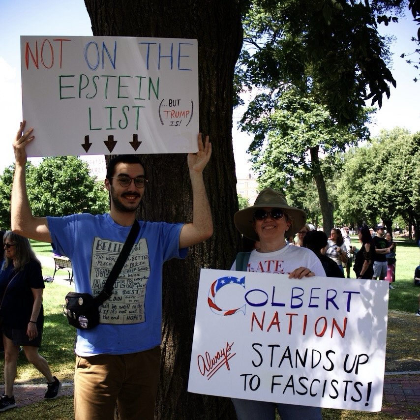 Photo of two people holding protest signs outdoors. One person is holding the sign above their head. Text reads “NOT ON THE EPSTEIN LIST (But Trump is)”. The other person’s sign says “COLBERT NATION ALWAYS STANDS UP TO FASCISTS”.