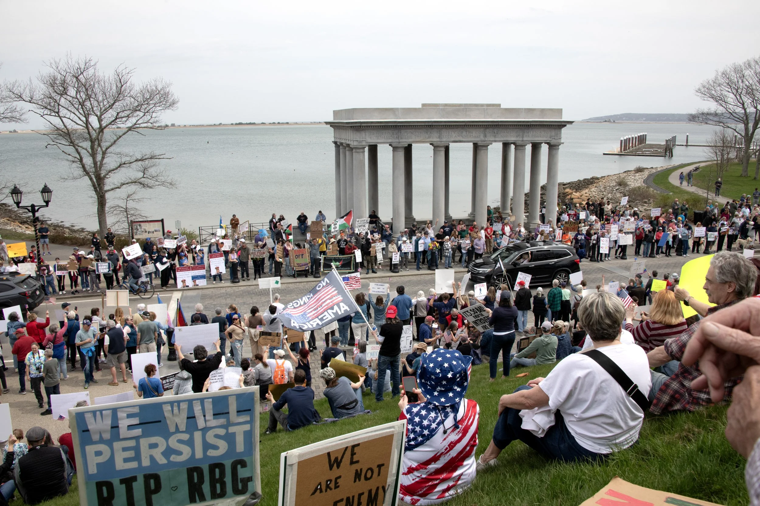 A protest taking place in Plymouth Hill Bay with the ocean in the background on a dreary spring day. There are signs saying "We Will Persist" and "No Kings in America"
