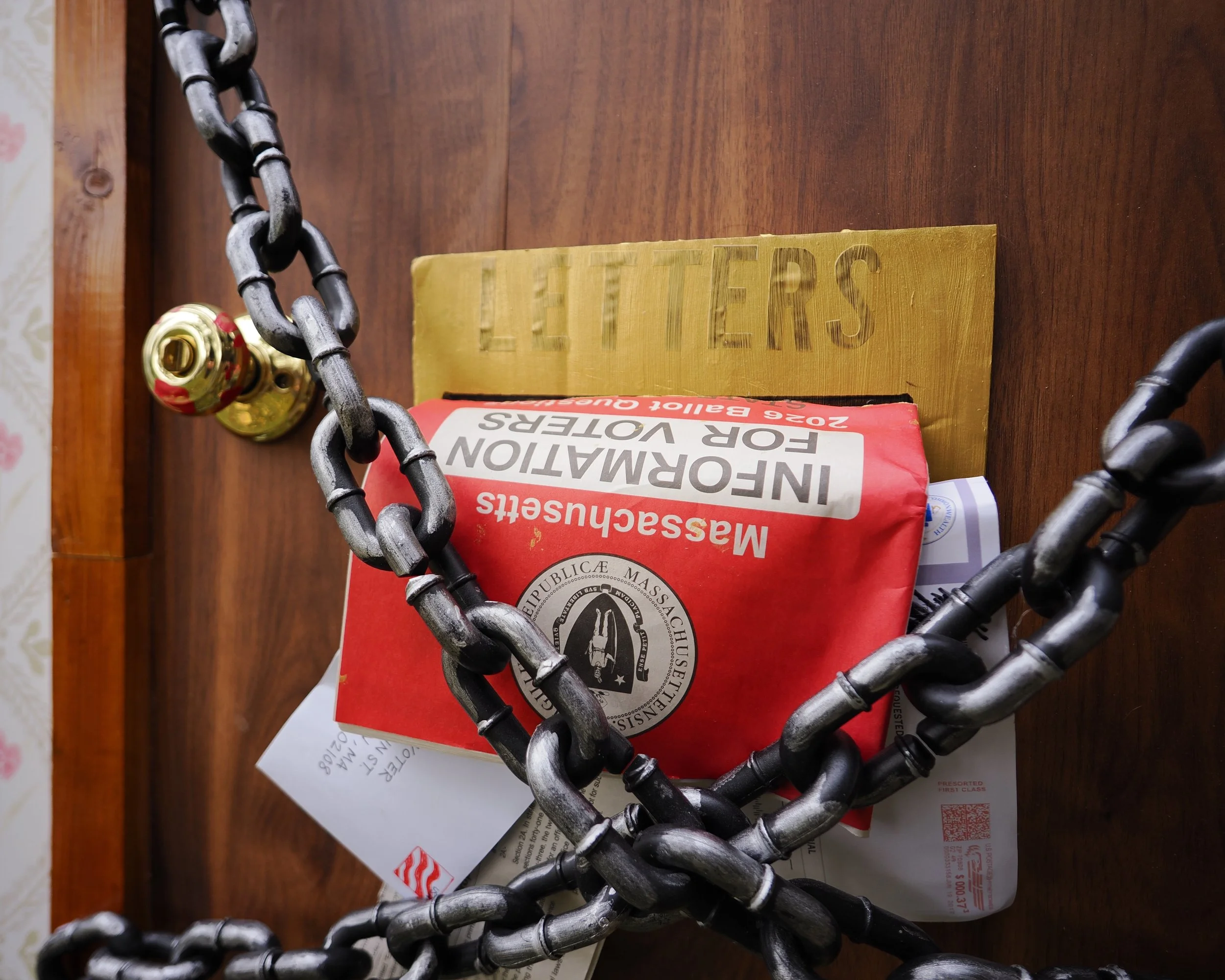 Chains over a door with a mail slot containing the Massachusetts Information for Voters guide and other letters.