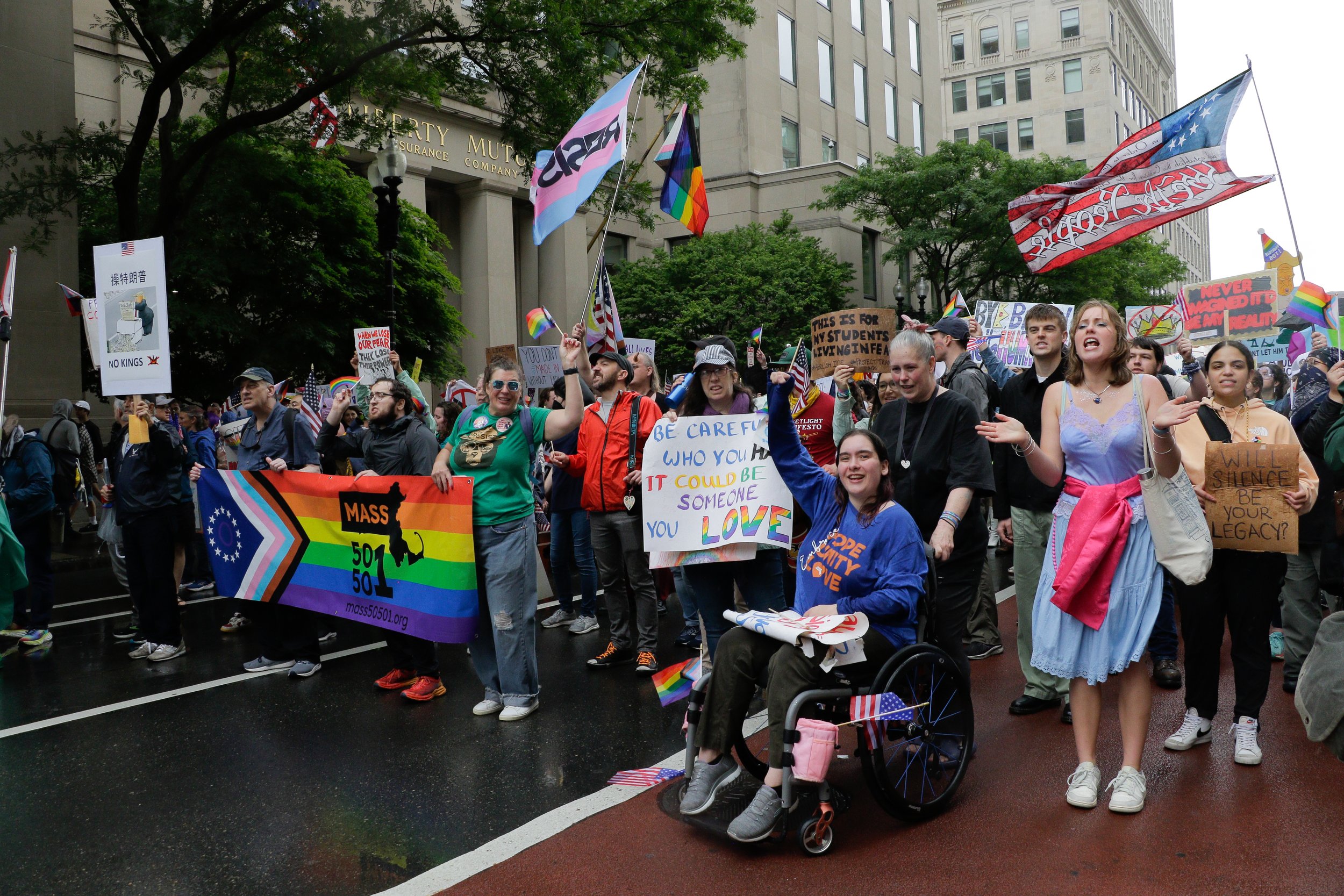Protestors marching behind a large progress pride Mass 50501 banner. Notable visuals include a sign saying "Will silence be your legacy", "this is for my students living in fear" and "be careful who you hate. it could be someone you love."