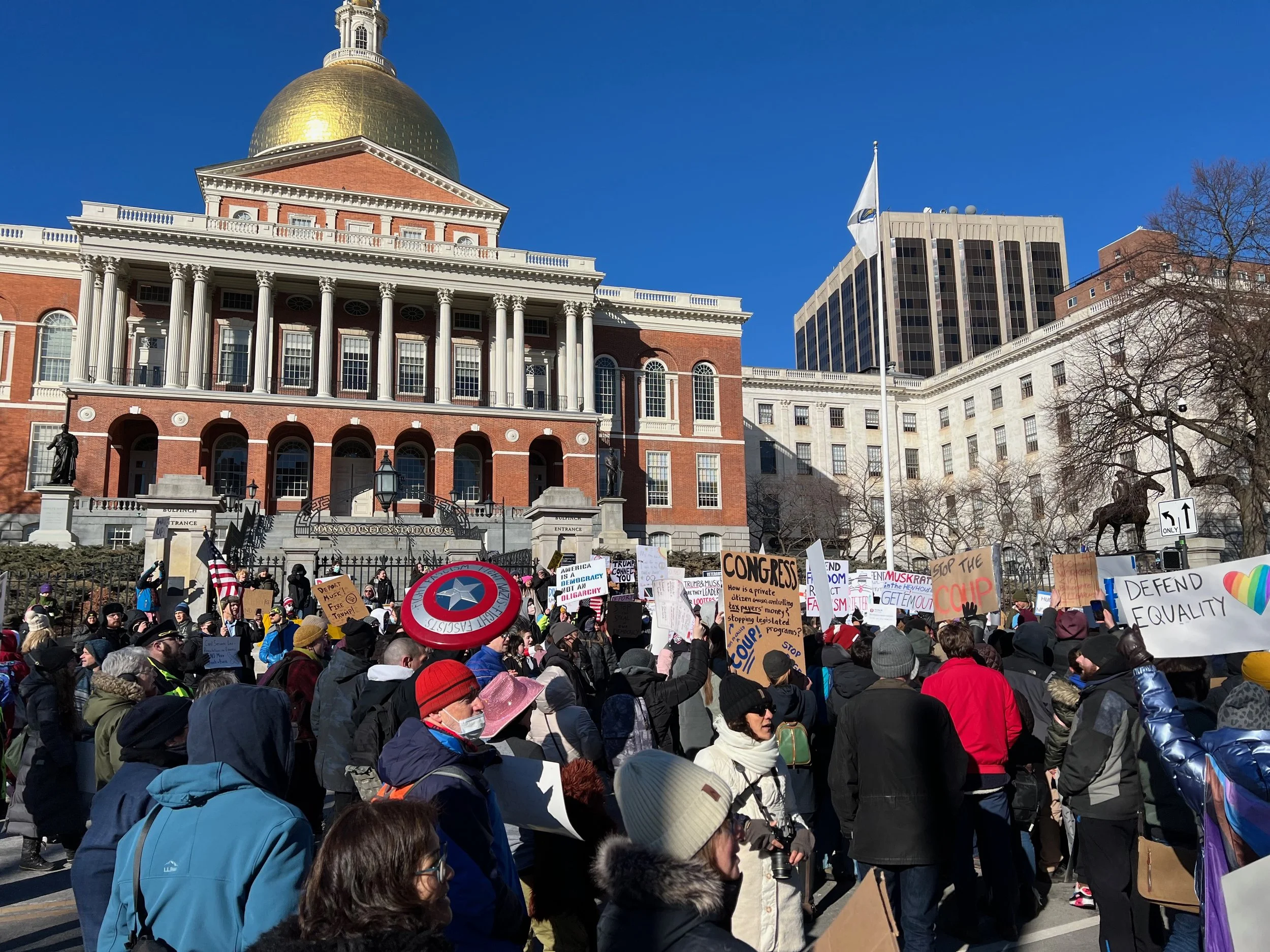 Picture of the first protest for 50501 in Boston. It shows the state house with hundreds of protestors and various signs, including Captain America's shield and one saying "Defend Equality" in focus.