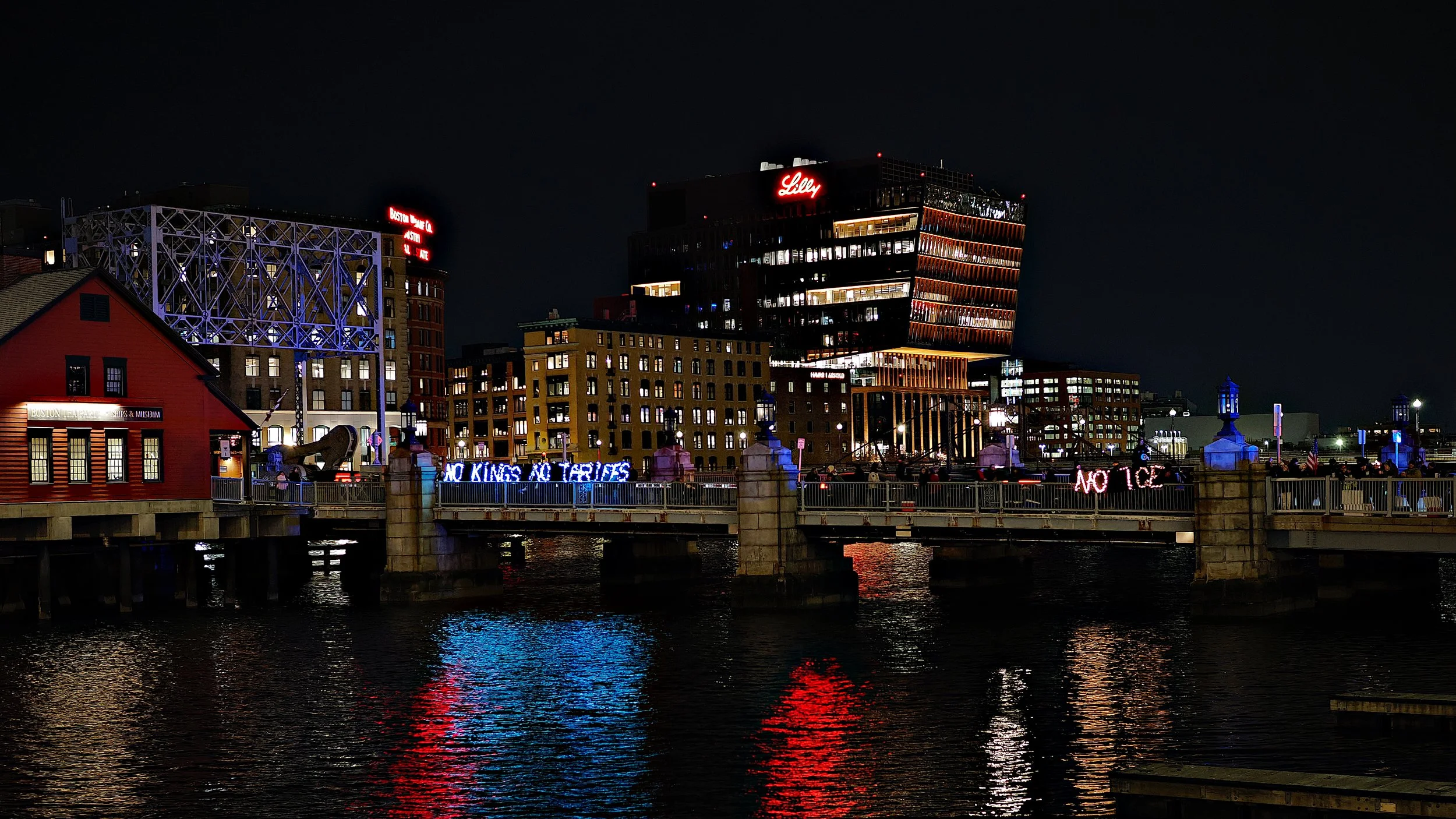 A nighttime photo of lights spelling out  “No Kings,” “No Tariffs,” and “No ICE” next to the Boston Tea Party museum.